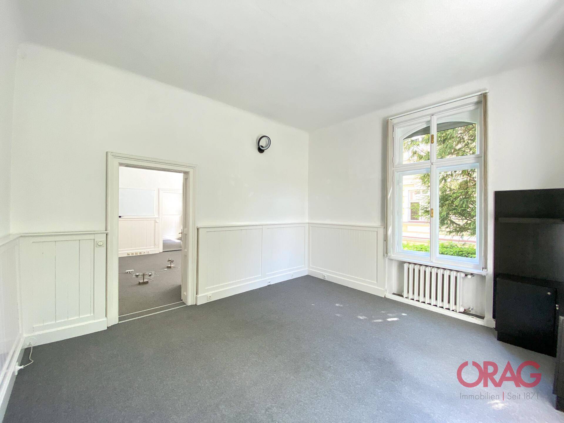 Helles Zimmer mit Teppichboden, einem großen Fenster und weißer Holzvertäfelung an den Wänden.