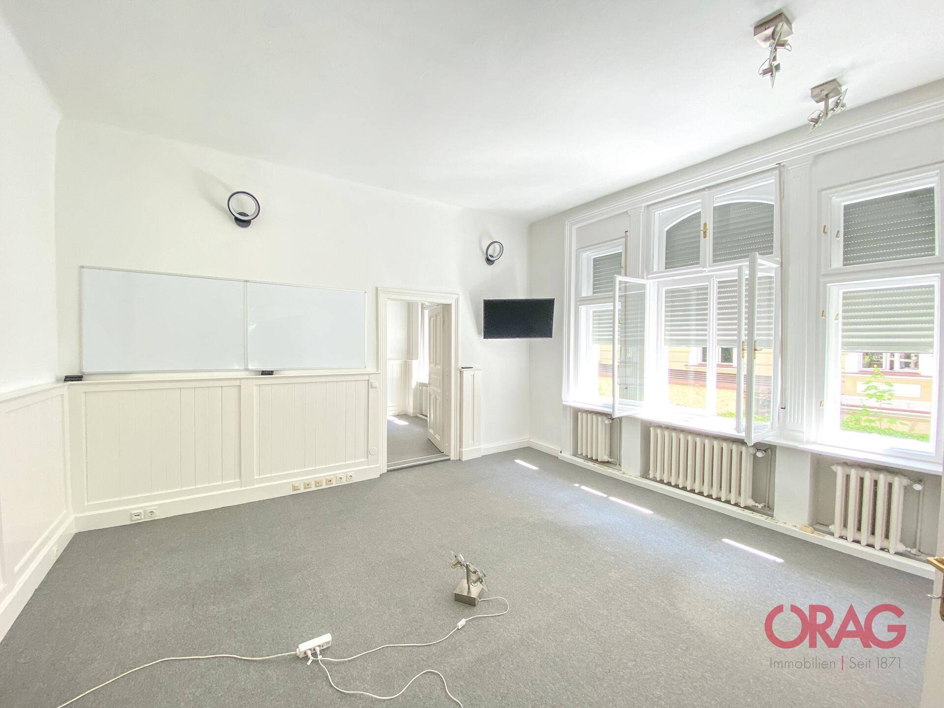Großer Raum mit Teppichboden, Fenstern mit Jalousien und einer Wandtafel für Präsentationen.