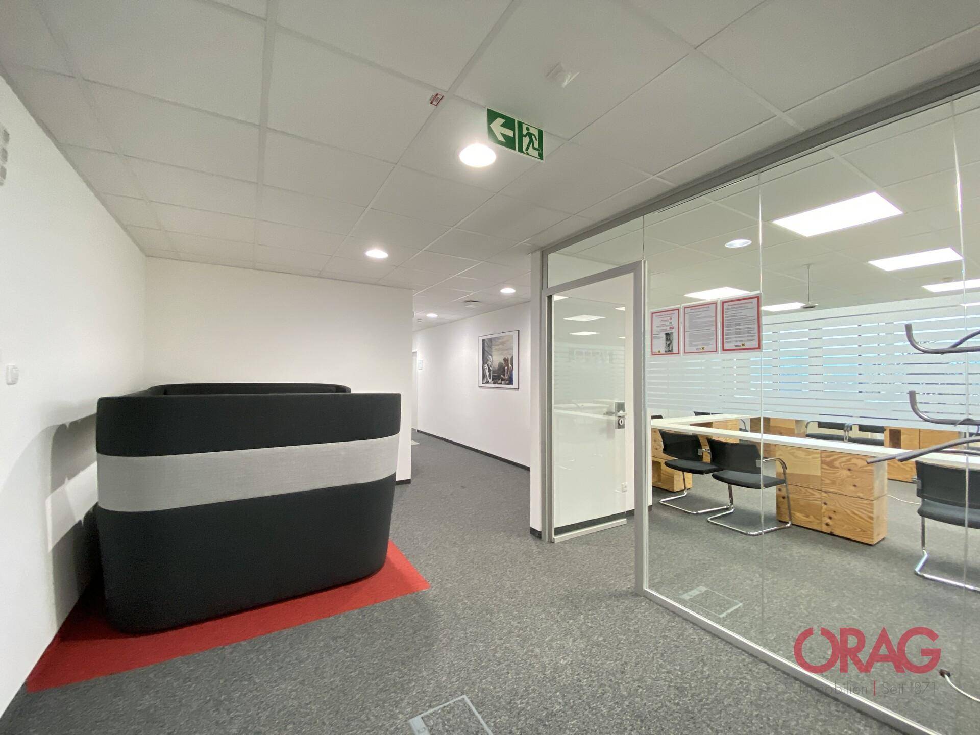 Heller Bürogang mit Teppichboden, Glaswänden und einem modernen Sitzmöbel.