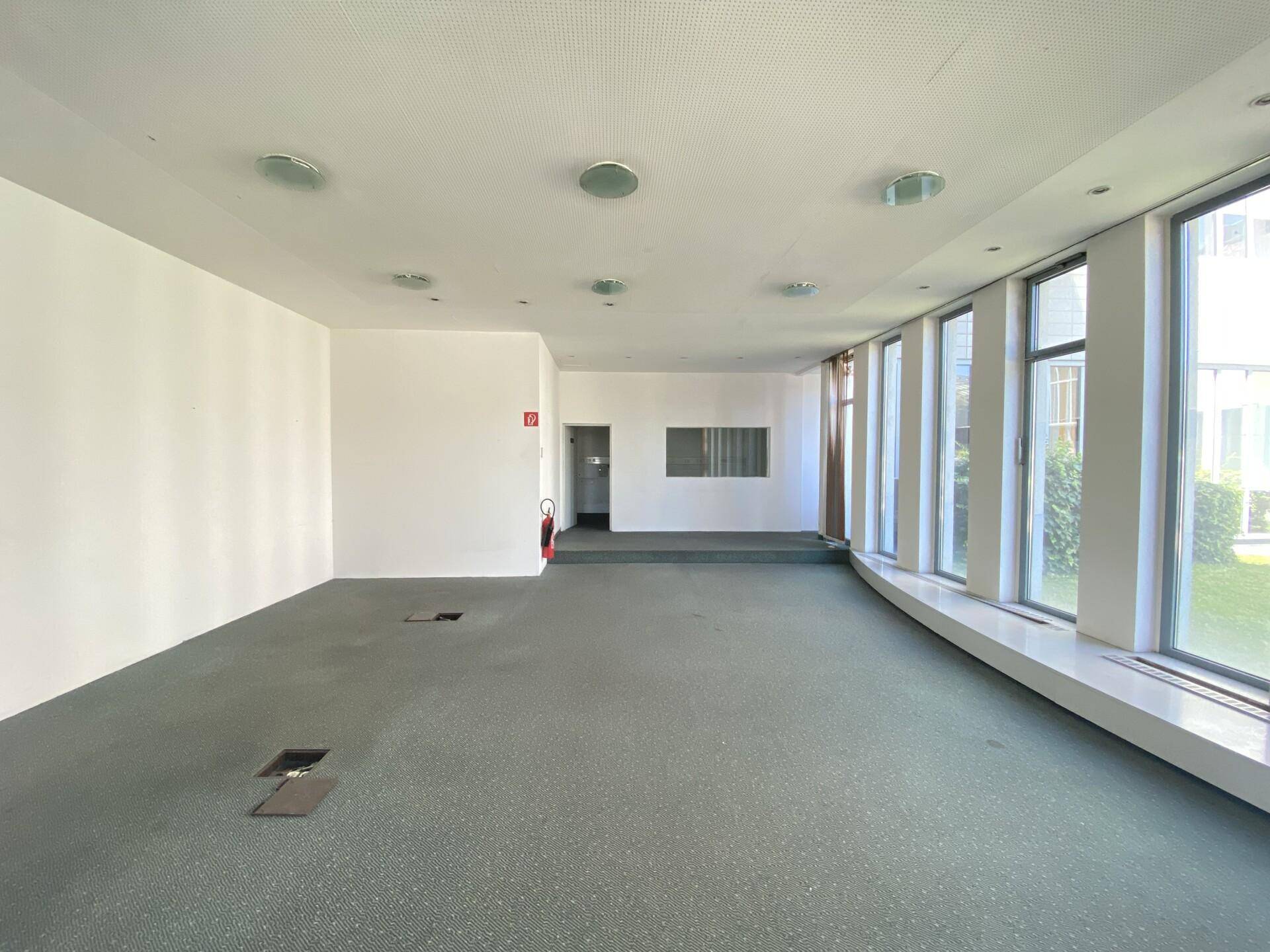 Großer, heller Büroraum mit Teppichboden und einer langen Fensterfront, die eine angenehme Arbeitsatmosphäre schafft.