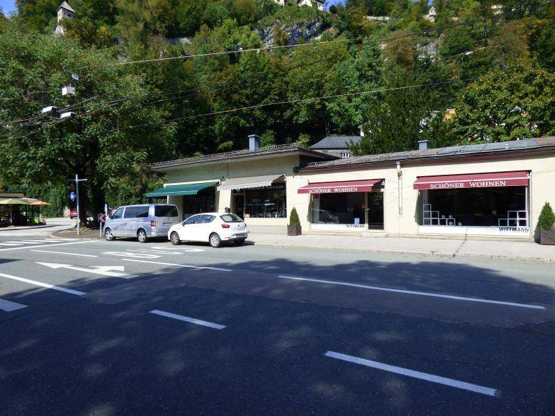 Straßenansicht des Geschäftshauses mit umliegenden Geschäften und bewaldetem Hügel im Hintergrund.