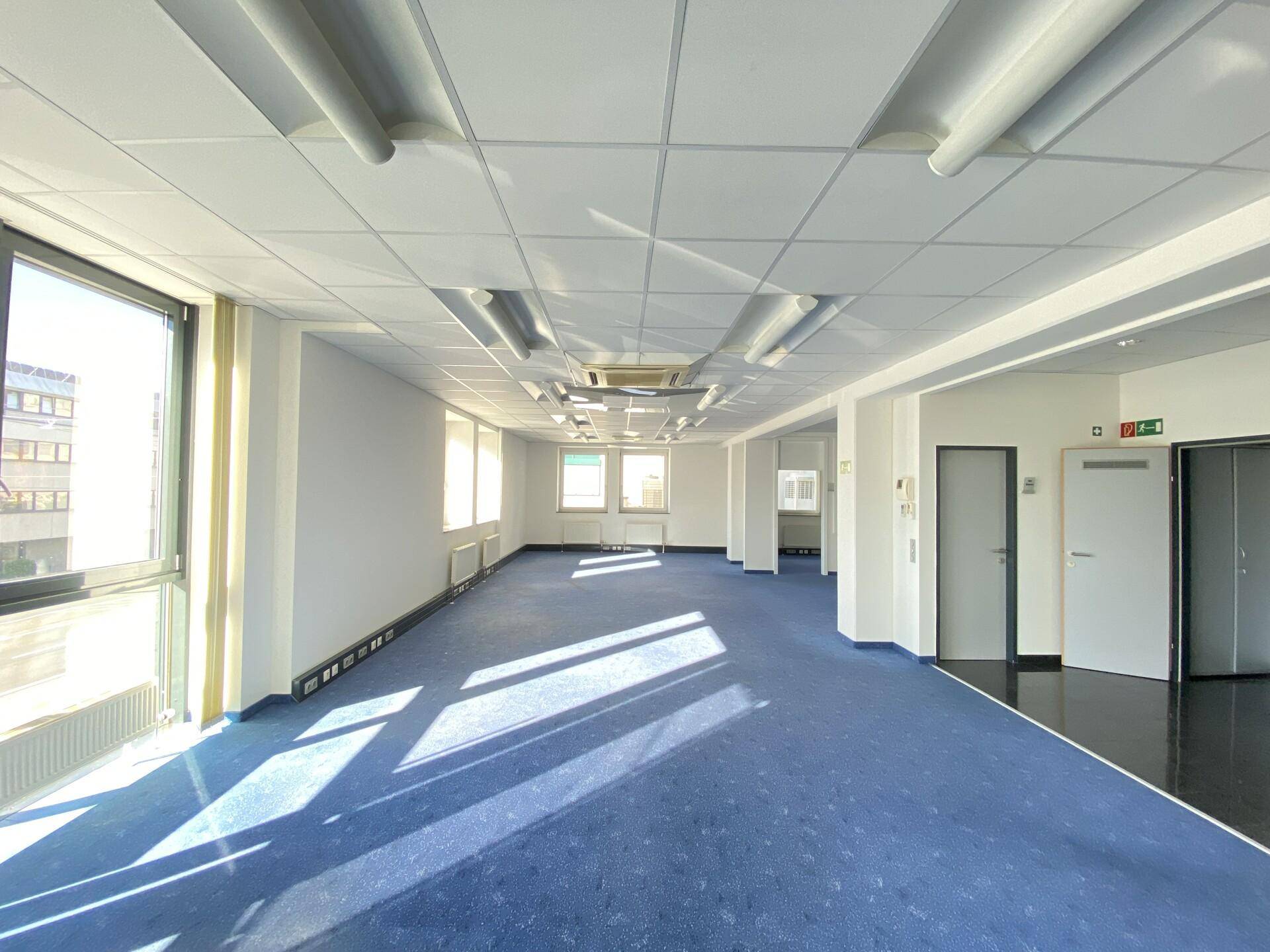 Geräumiges, leeres Büro mit blauem Teppichboden, Rasterdecke und großen Fenstern mit Bergblick.