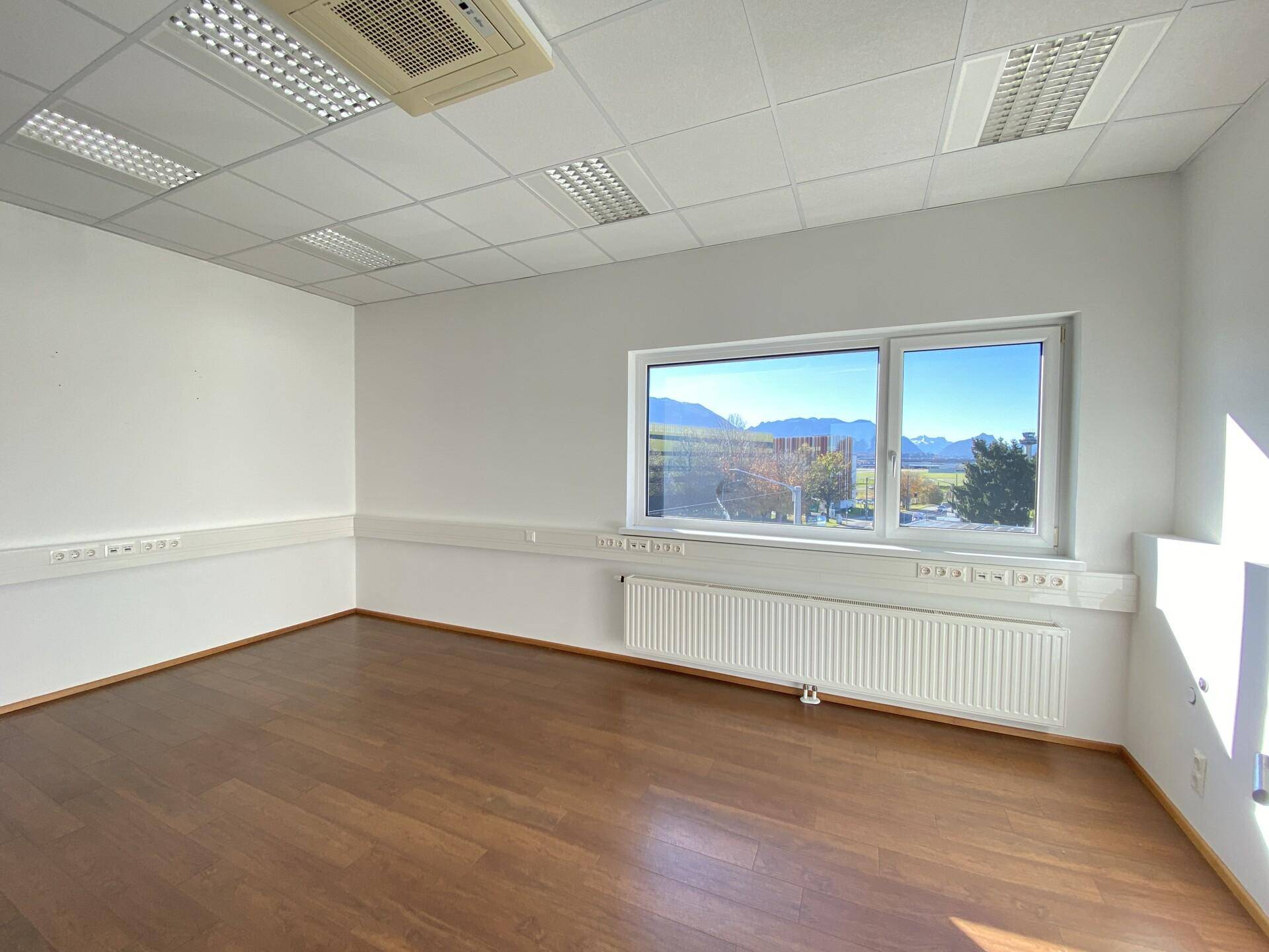 Leeres Büro mit Holzboden, weißen Wänden und Fenstern, die einen weiten Blick auf die Landschaft und Berge bieten.