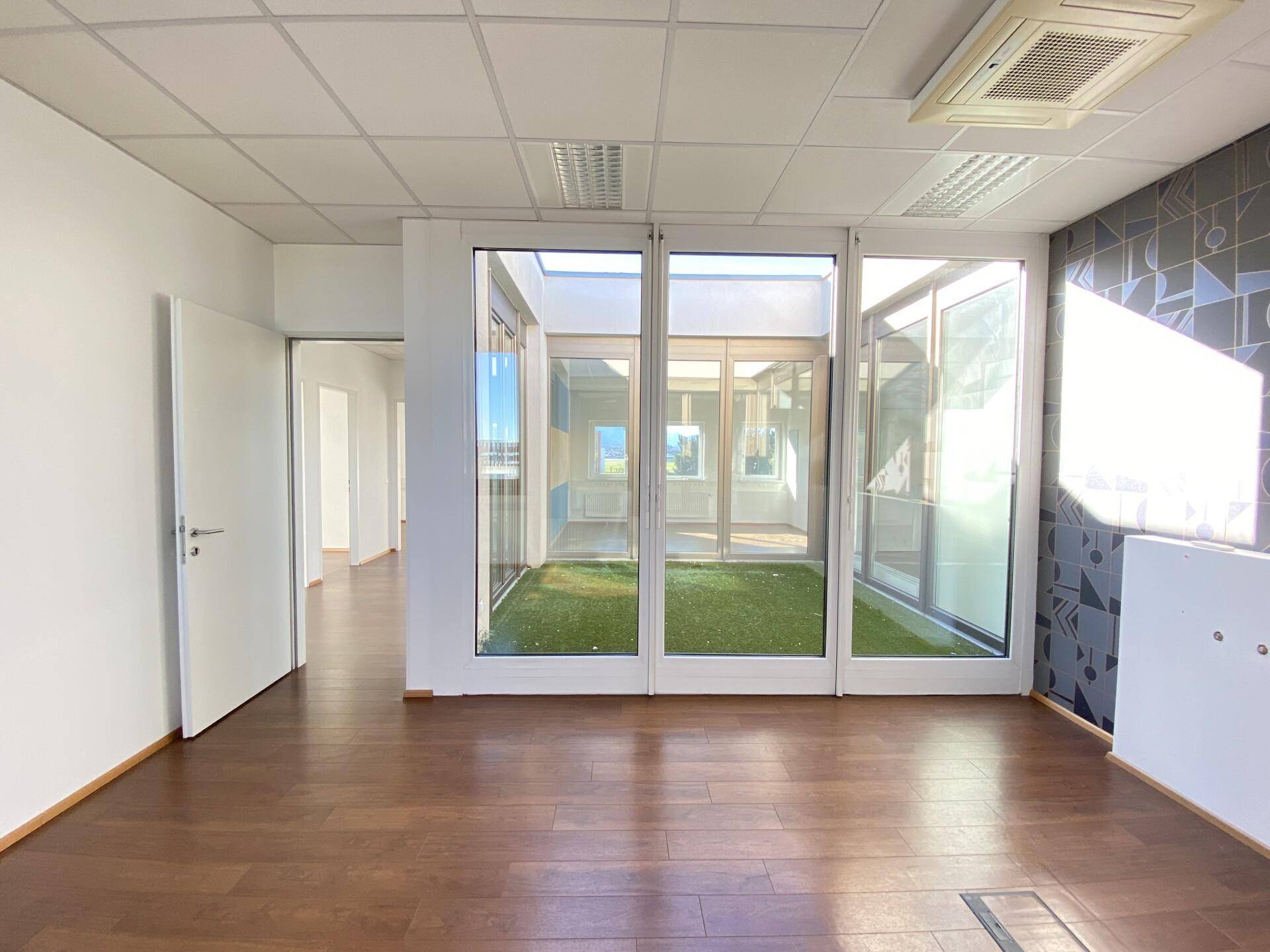 Modernes Büro mit Holzboden und Glaswänden, die einen Blick in ein sonniges, begrüntes Atrium ermöglichen.