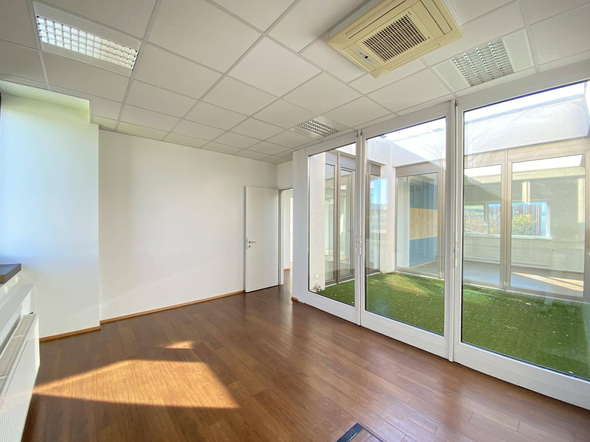 Geräumiges Büro mit Holzboden und großen Glasfronten, die einen Blick auf ein begrüntes Atrium bieten.