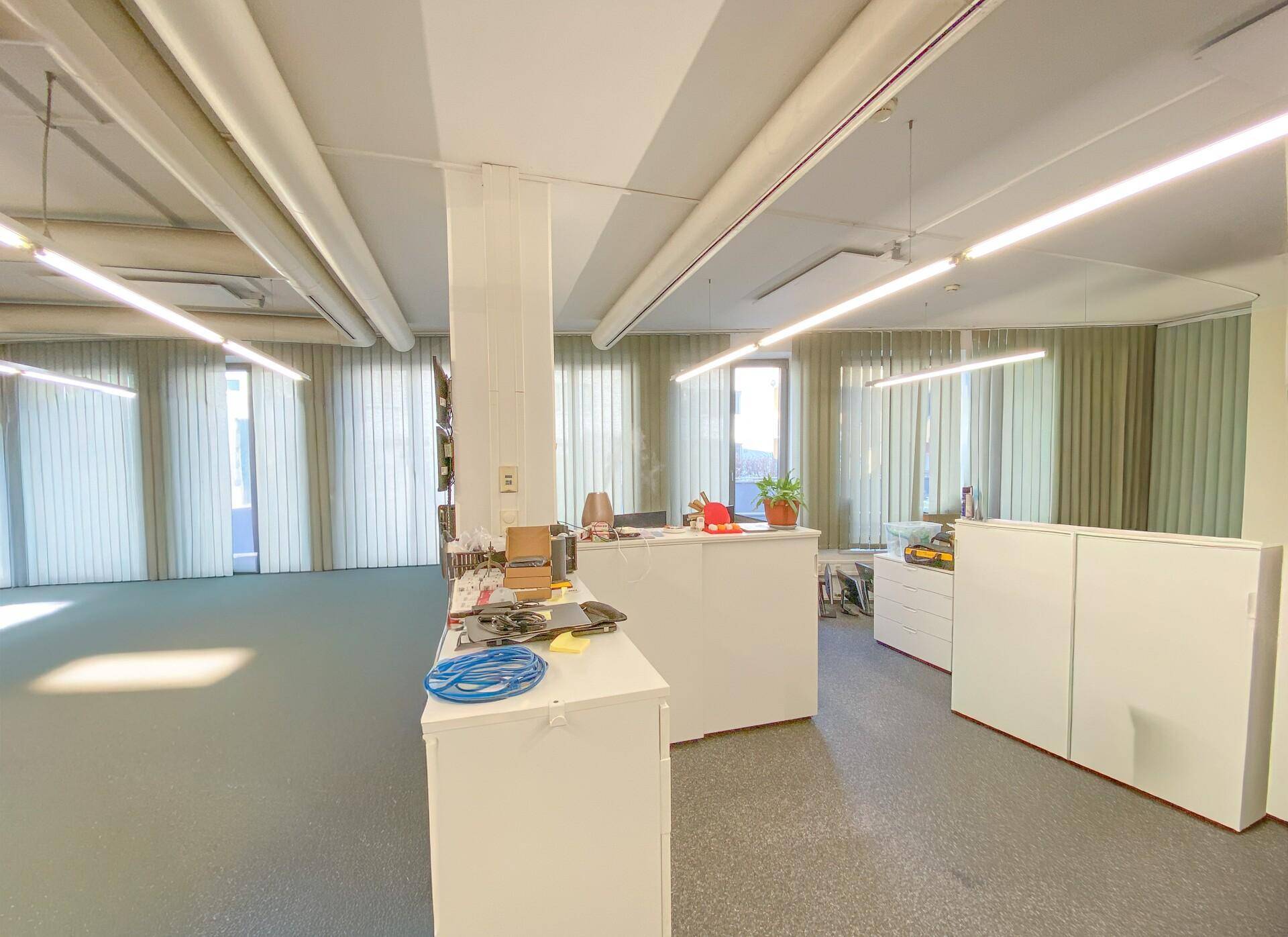 Modernes Büro mit mehreren Arbeitsplätzen, Säulen und großen Fenstern, die viel Tageslicht hereinlassen.