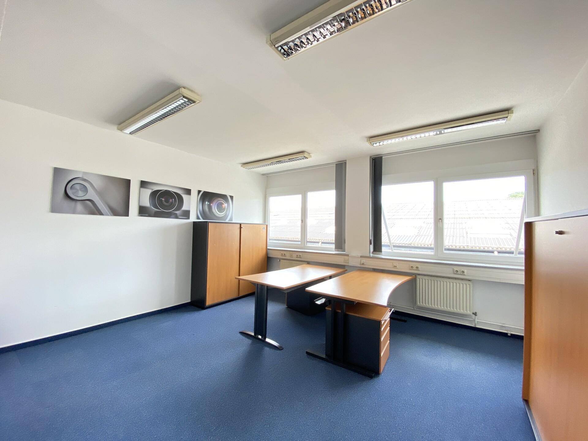 Büroraum mit blauer Teppichboden, Schreibtischen und Holzschränken, gut beleuchtet durch Fenster.