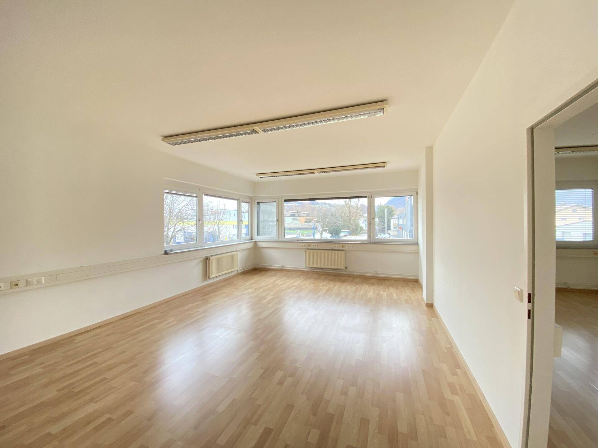 Geräumiger Büroraum mit hellem Parkettboden und einer Fensterfront, die viel Tageslicht hereinlässt.
