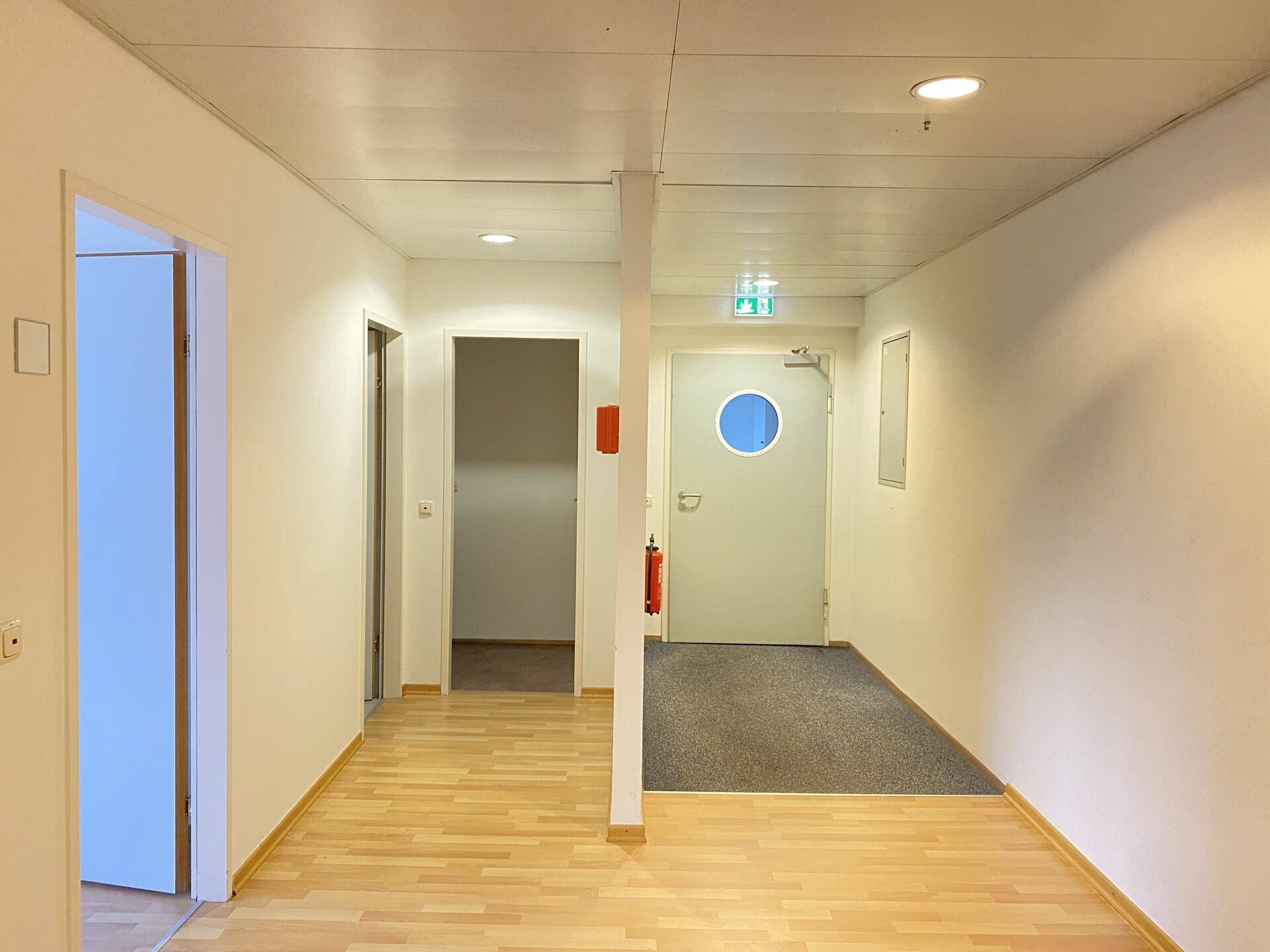 Heller Flur mit Holzboden, Brandschutzvorrichtungen und einem Ausgang mit runder Verglasung in der Tür.