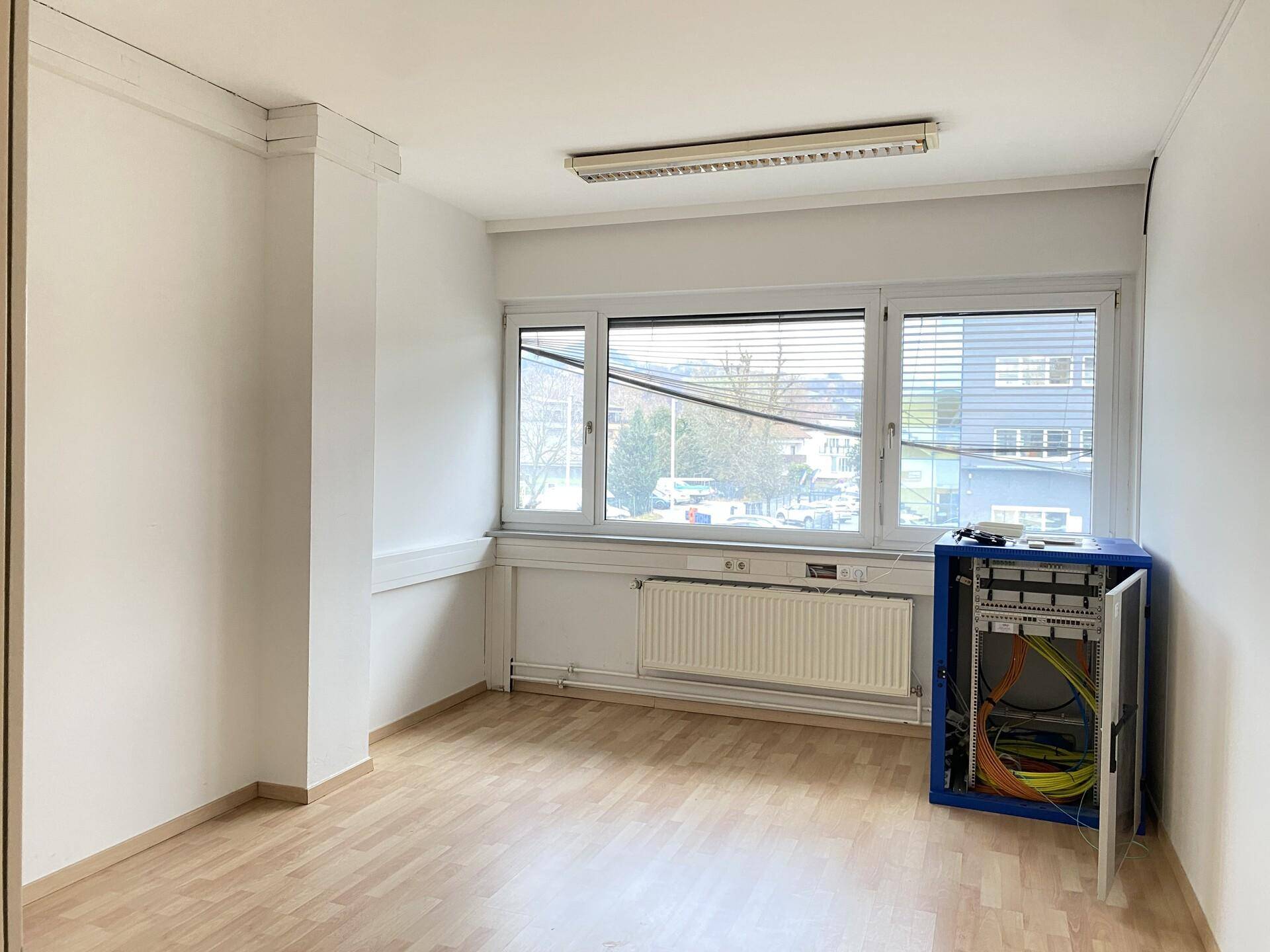 Büroraum mit großen Fenstern, Heizkörpern und einem offenen Netzwerkschrank an der Wand.