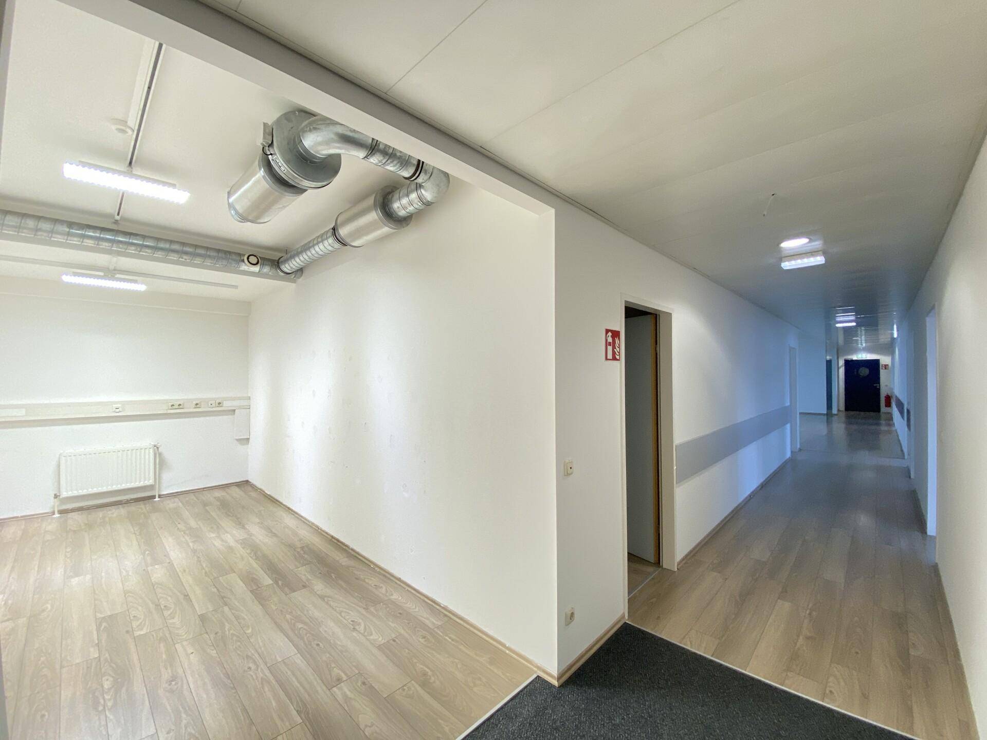 Gewerberaum mit sichtbaren Lüftungsrohren an der Decke, hellem Holzfußboden und Zugang zu einem langen Flur.