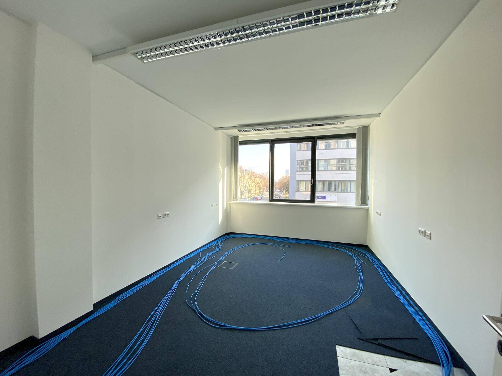 Kleinerer Büroraum mit blauem Teppichboden und verlegten Kabeln, bereit für die Einrichtung.