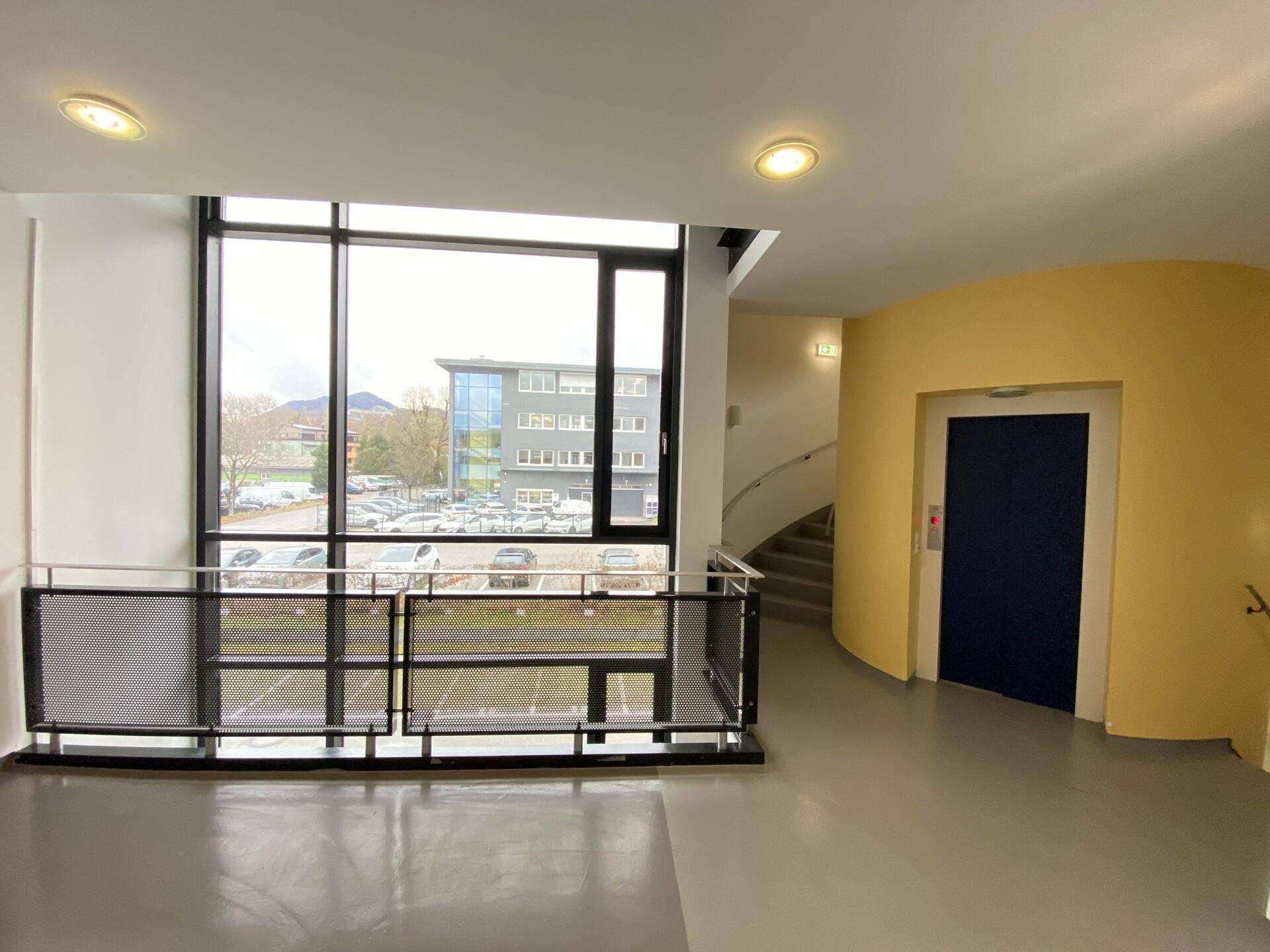 Heller Eingangsbereich mit Aufzug und Wendeltreppe, großen Fenstern und Blick auf den Parkplatz.