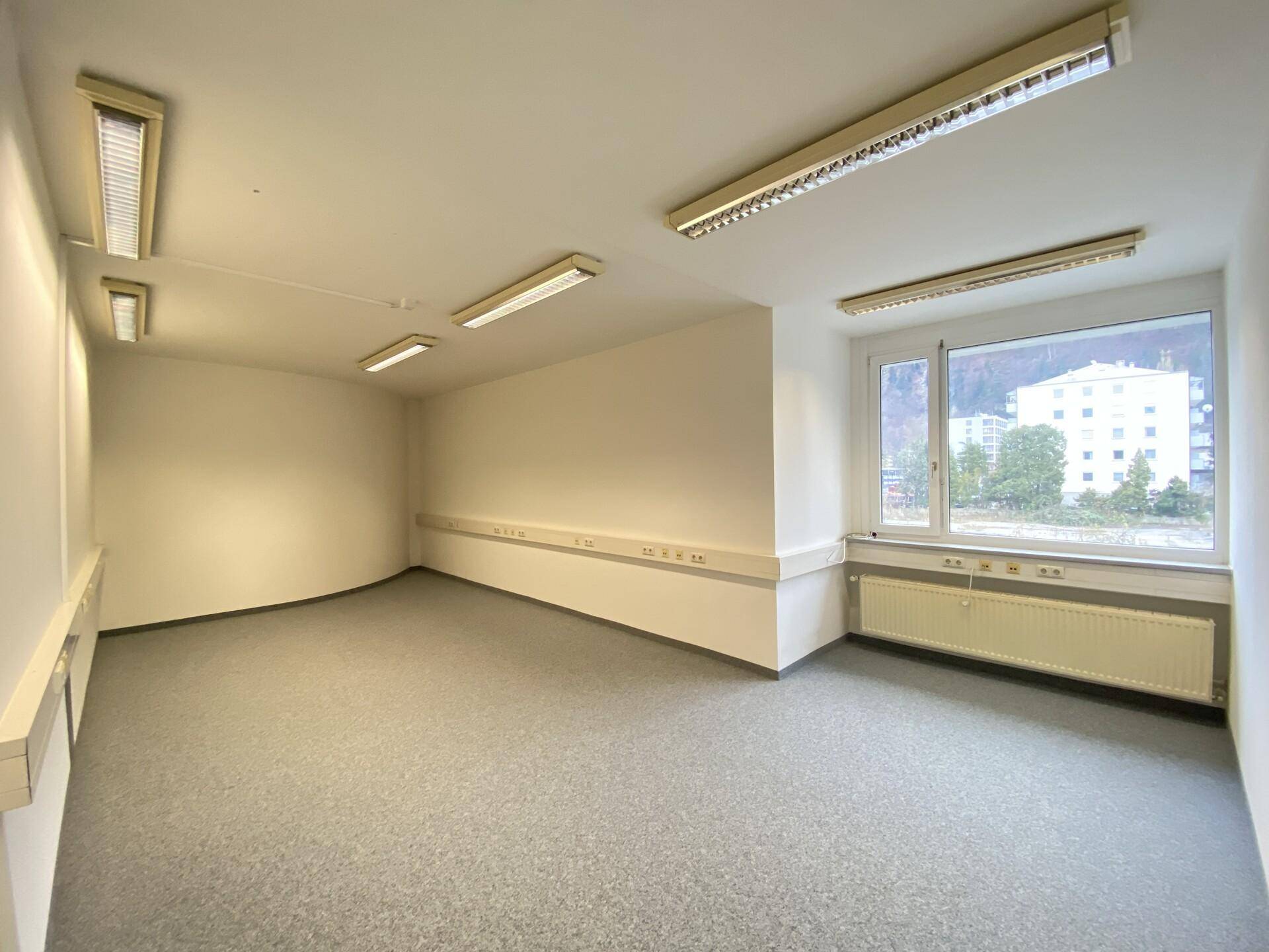 Helles Büro mit Teppichboden und großen Fenstern, die einen Blick auf die Umgebung ermöglichen.