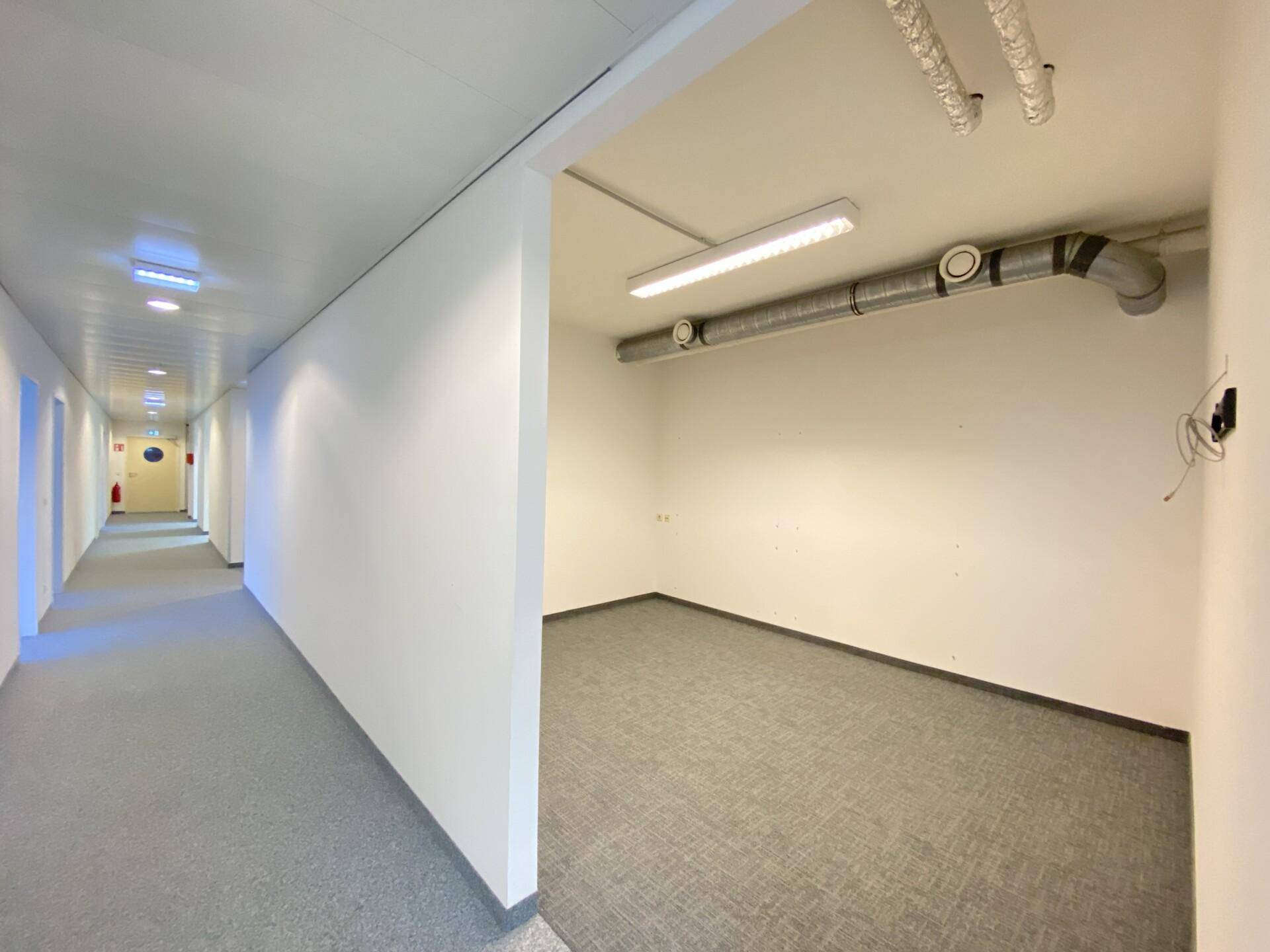 Moderner Flur mit Teppichboden und sichtbaren Lüftungsrohren, der zu weiteren Büroräumen führt.