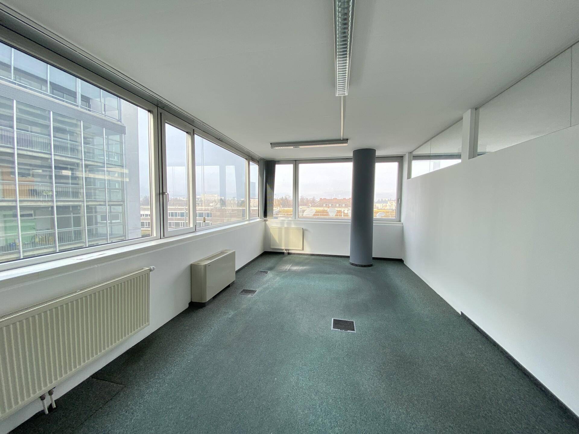 Helles Eckbüro mit großen Fenstern und Teppichboden, bietet einen Panoramablick auf die Stadt.