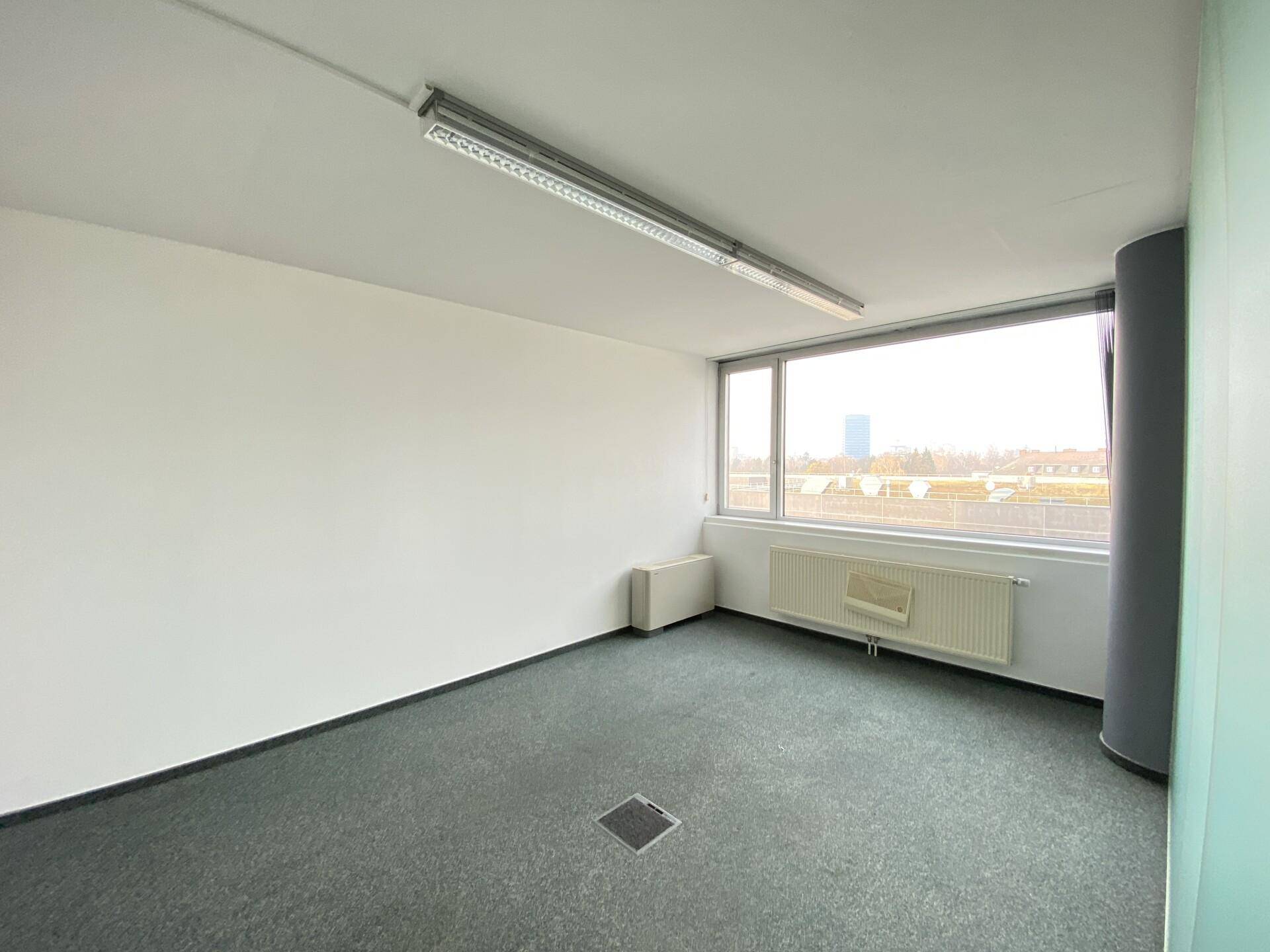 Einfacher Büroraum mit Teppichboden und einem großen Fenster, bietet ausreichend Tageslicht.