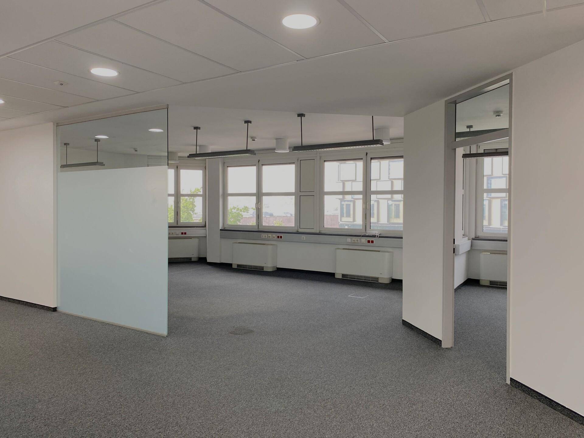 Geräumiges Büro mit grauem Teppichboden, großen Fenstern und einer modernen Glaswand.