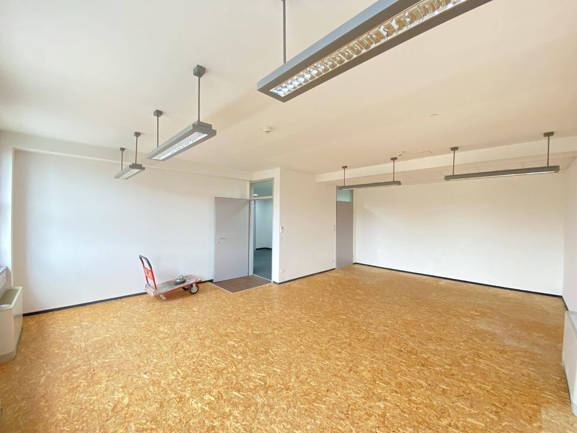 Leeres Büro mit OSB-Plattenboden, weißen Wänden und modernen Deckenleuchten.