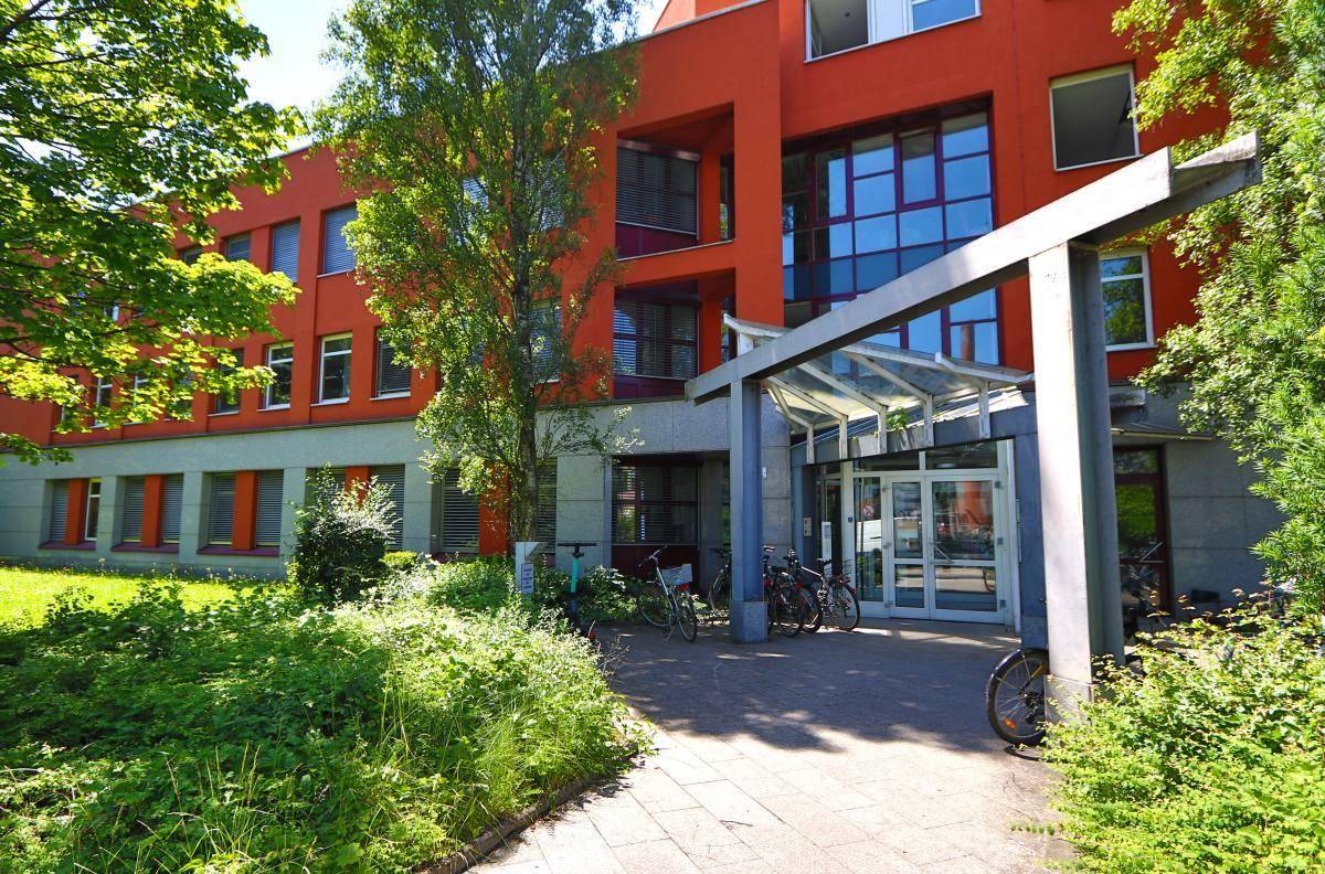 Modernes Bürogebäude mit roter Fassade, großen Fenstern und begrüntem Eingangsbereich.