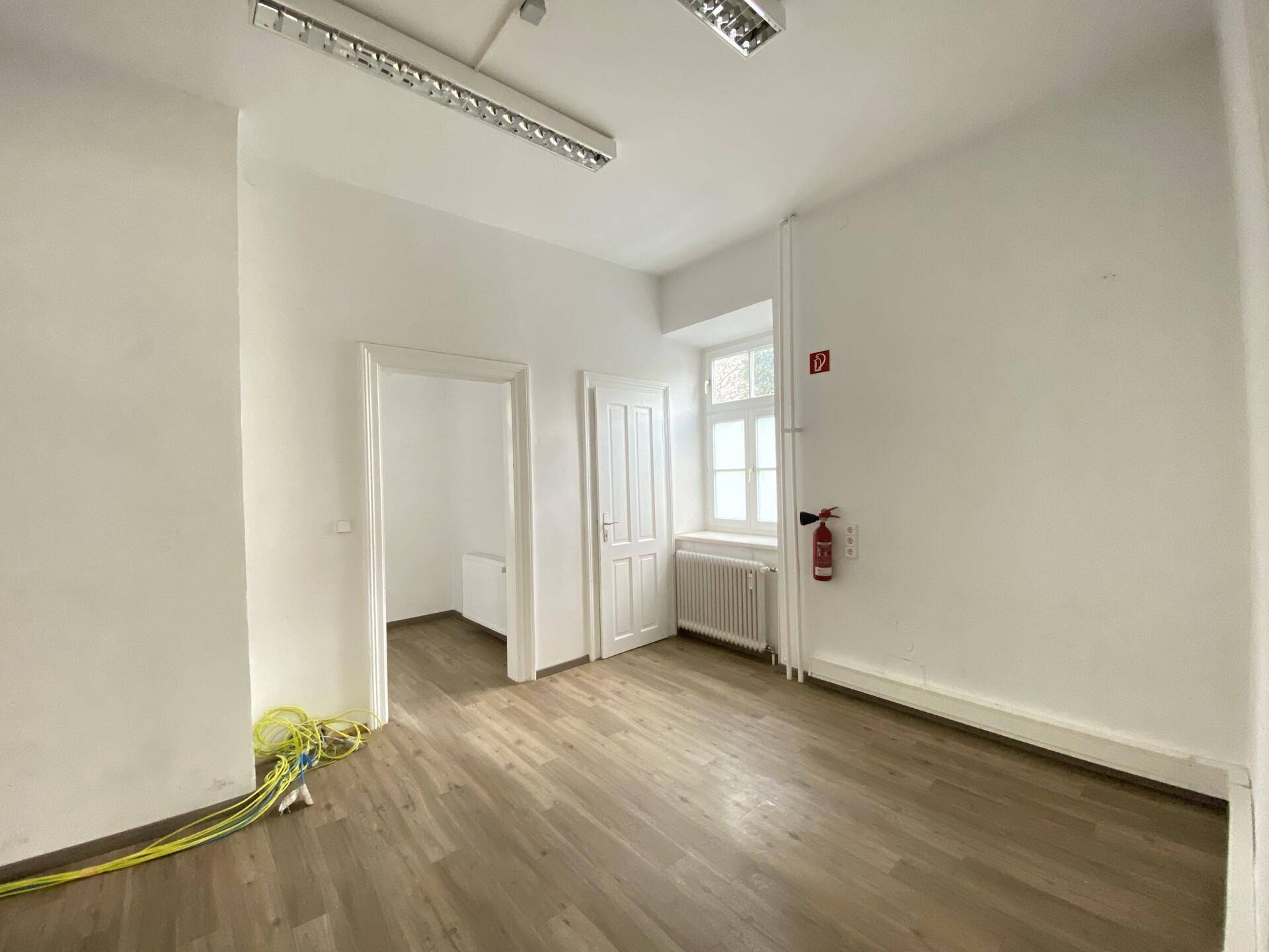 Kleinerer Büroraum mit Laminatboden, Fenster und Deckenbeleuchtung, inklusive Feuerlöscher.