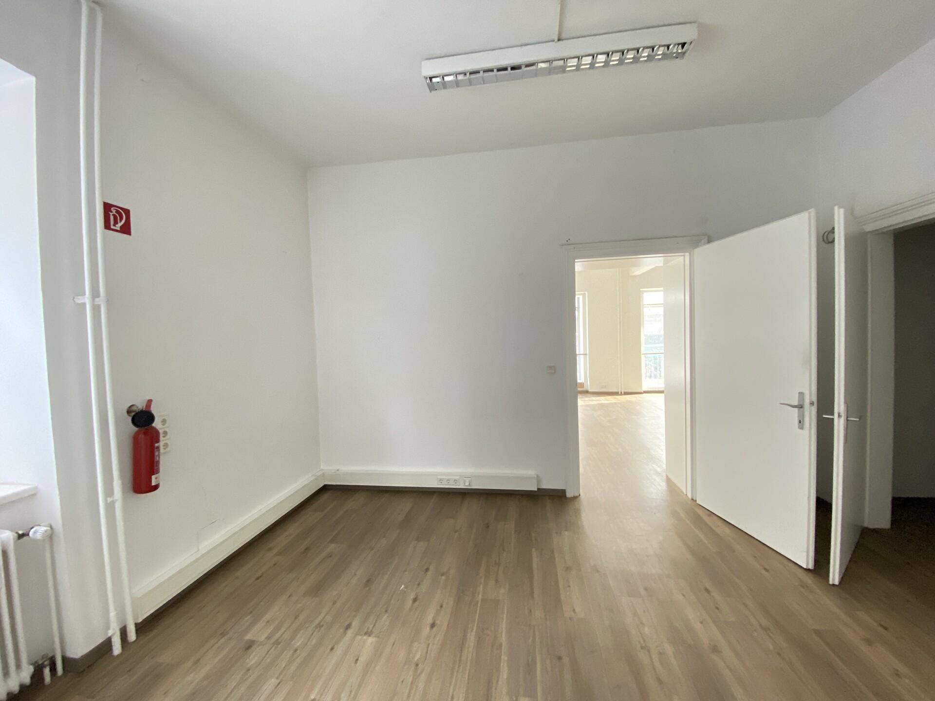 Heller Büroraum mit Laminatboden und Zugang zu weiteren Bereichen, gut beleuchtet.