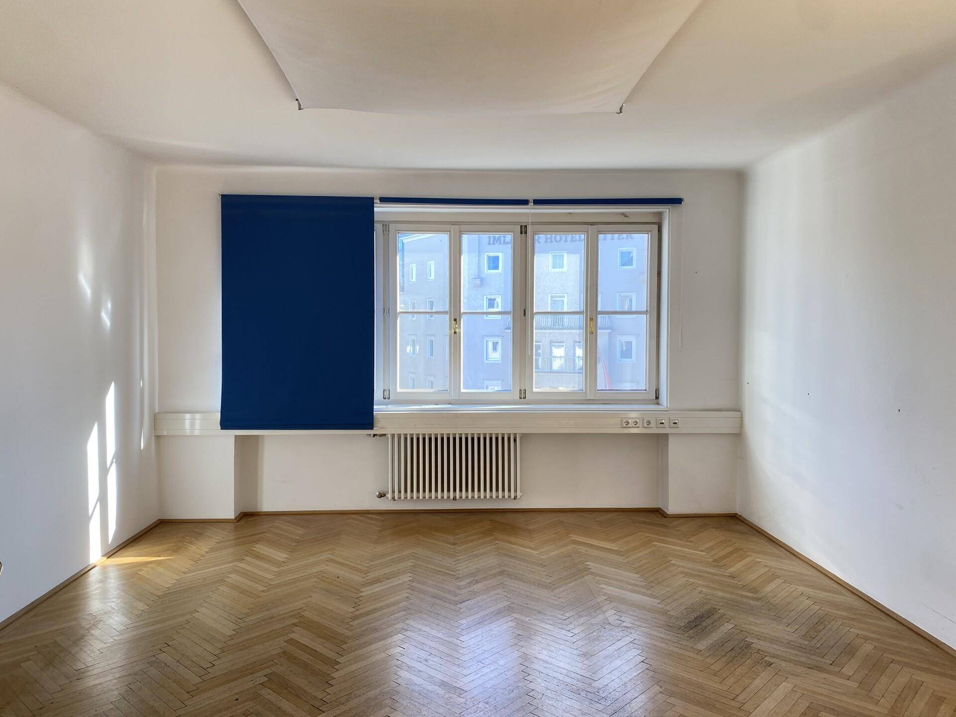 Heller Büroraum mit Fischgrätparkett, großem Fenster und blauem Rollo, das die Aussicht verdeckt.