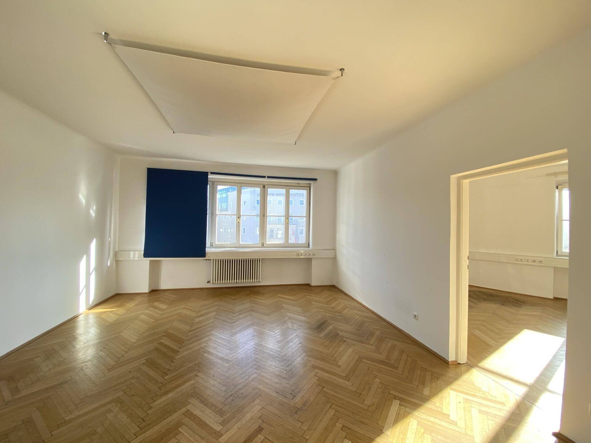 Lichtdurchfluteter Bürobereich mit Fischgrätparkett und blauen Rollos an den Fenstern, die die Stadt zeigen.
