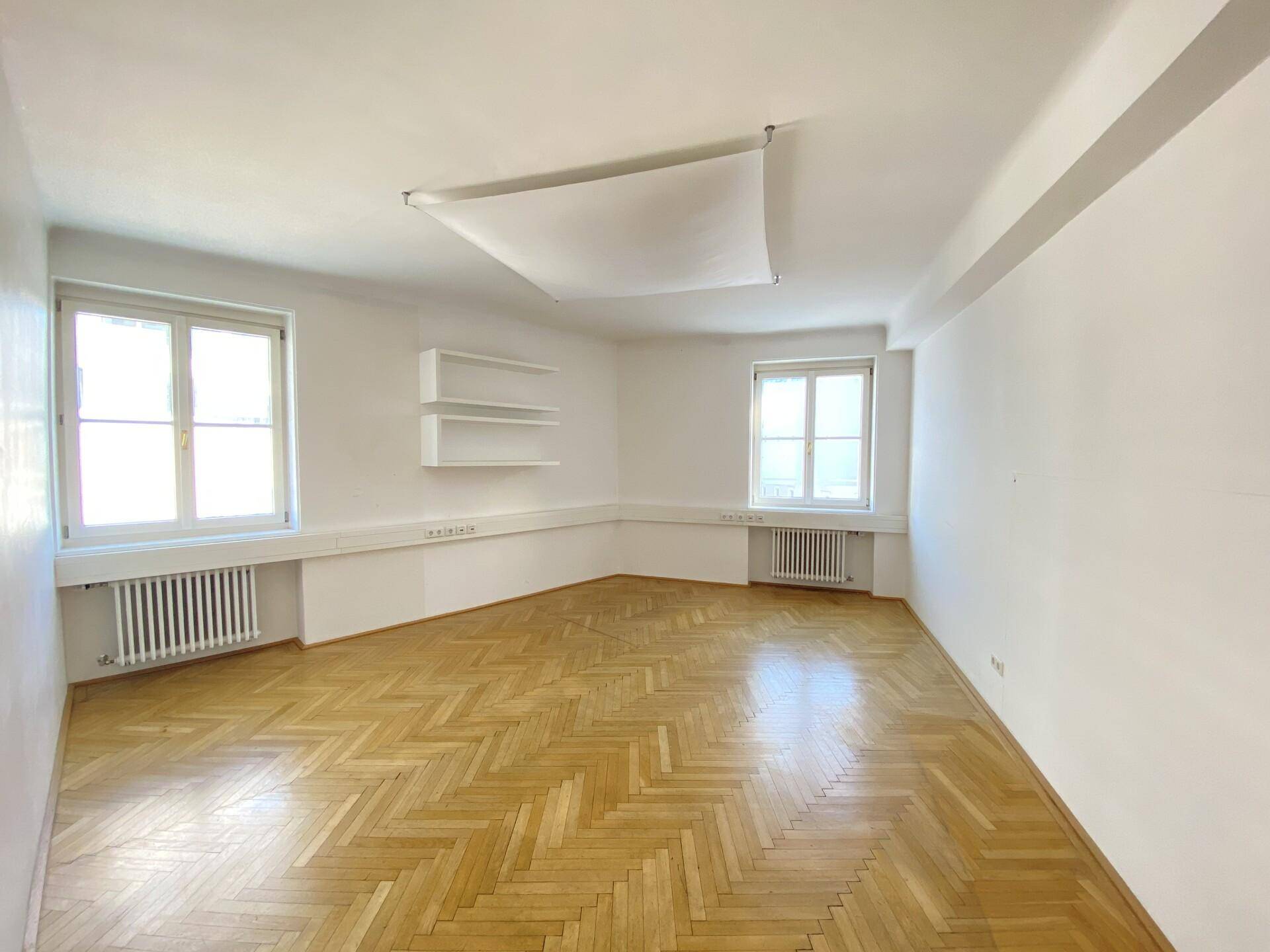 Heller Büroraum mit zwei Fenstern, Holzparkett und weißen Wandregalen für Ablageflächen.