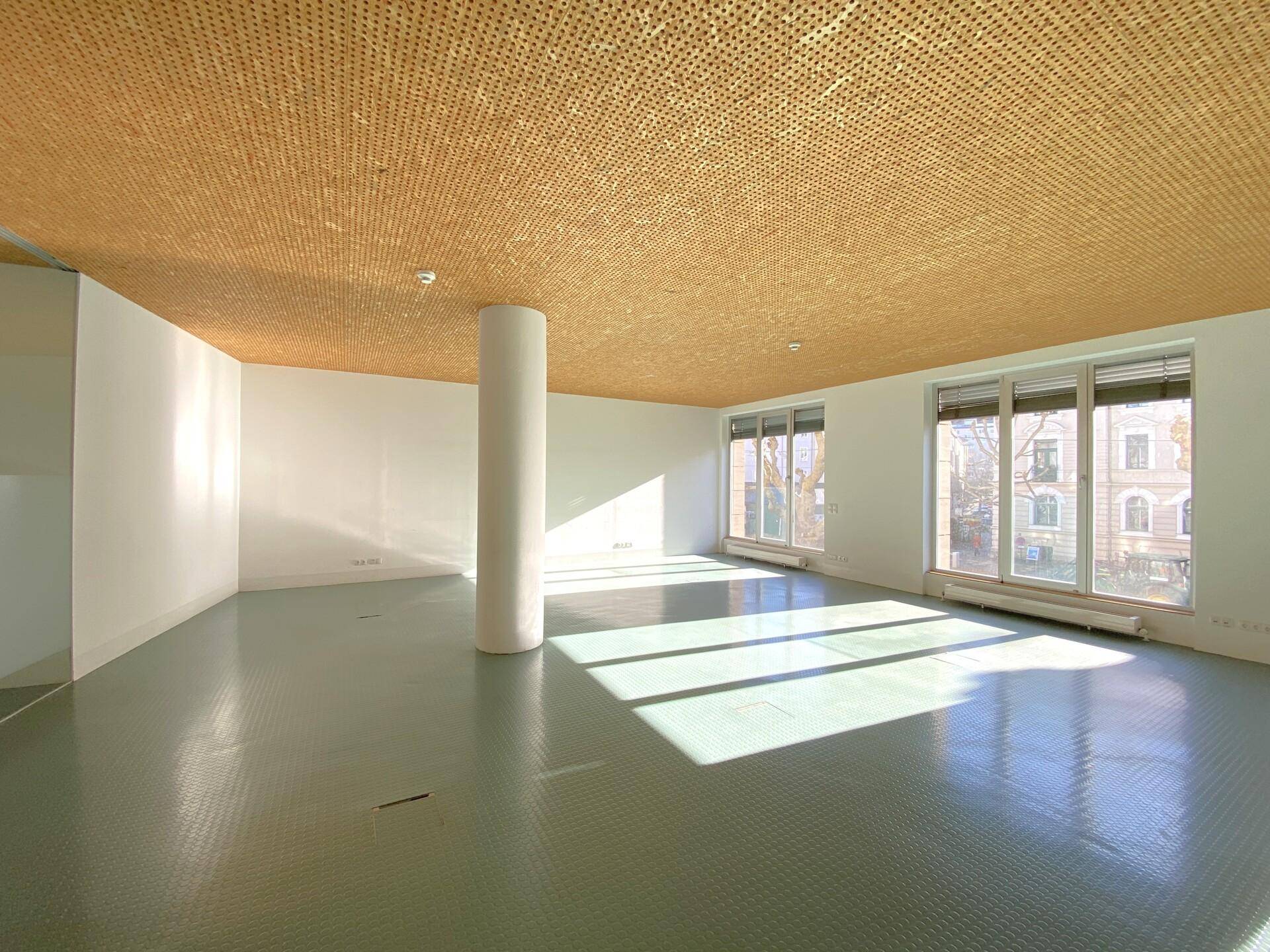 Heller Büroraum mit großen Fenstern, die viel Sonnenlicht hereinlassen, und einer markanten Holzdecke.