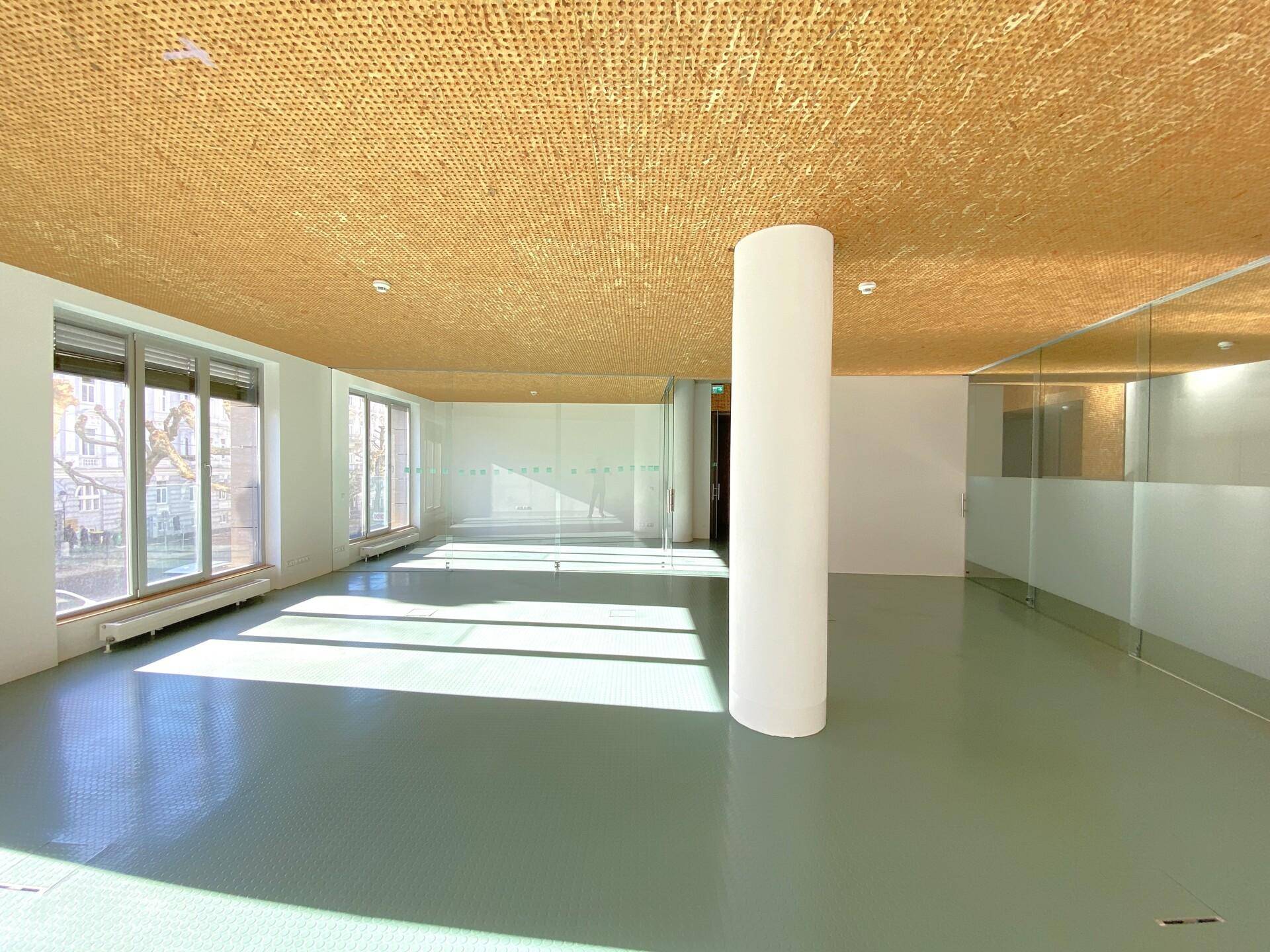 Geräumiger Bürobereich mit einer zentralen Säule, großen Fenstern und einer akustischen Holzdecke.