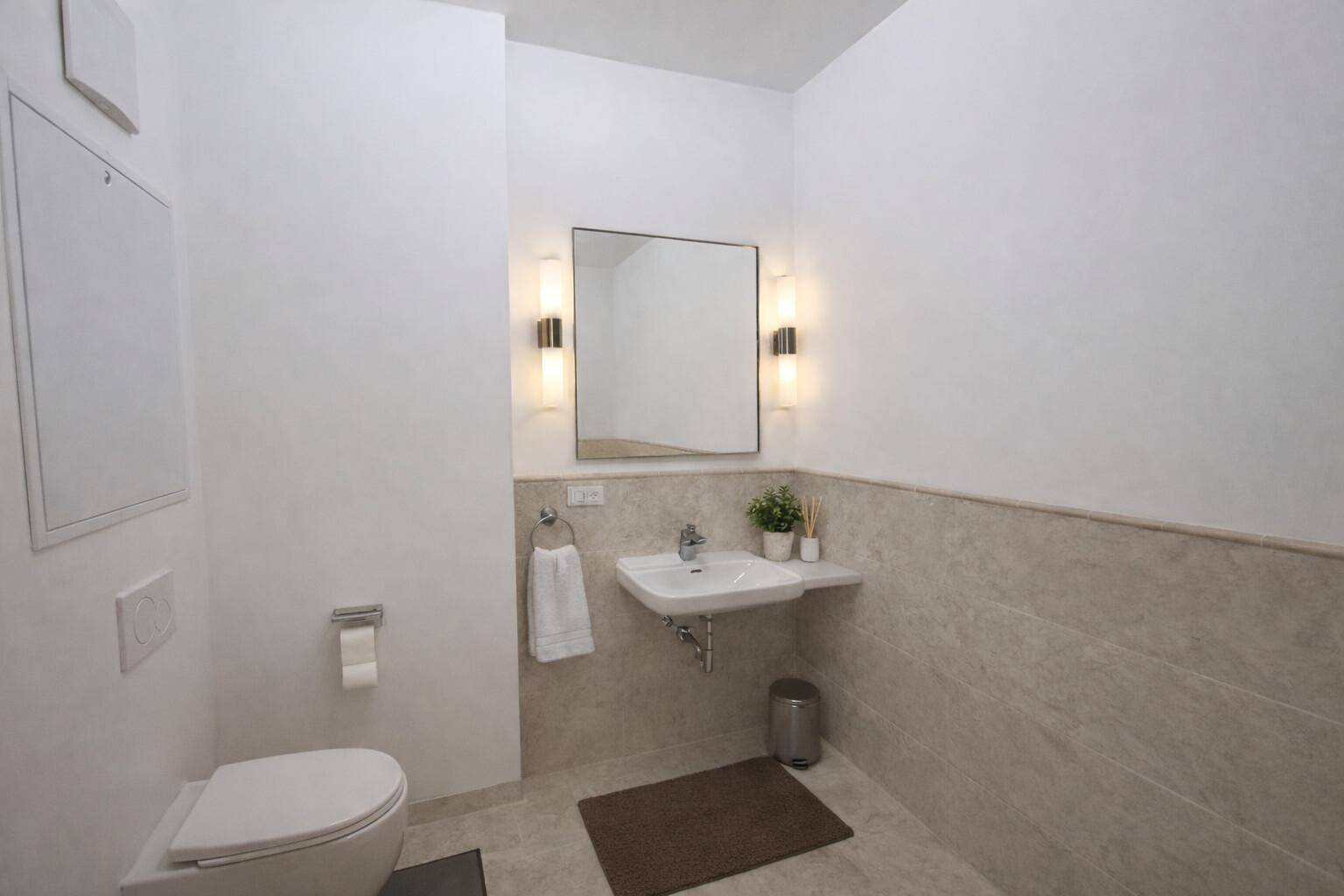 Modernes Badezimmer mit wandhängender Toilette, Waschbecken und großem Spiegel, teilweise gefliest mit hellen Steinplatten.