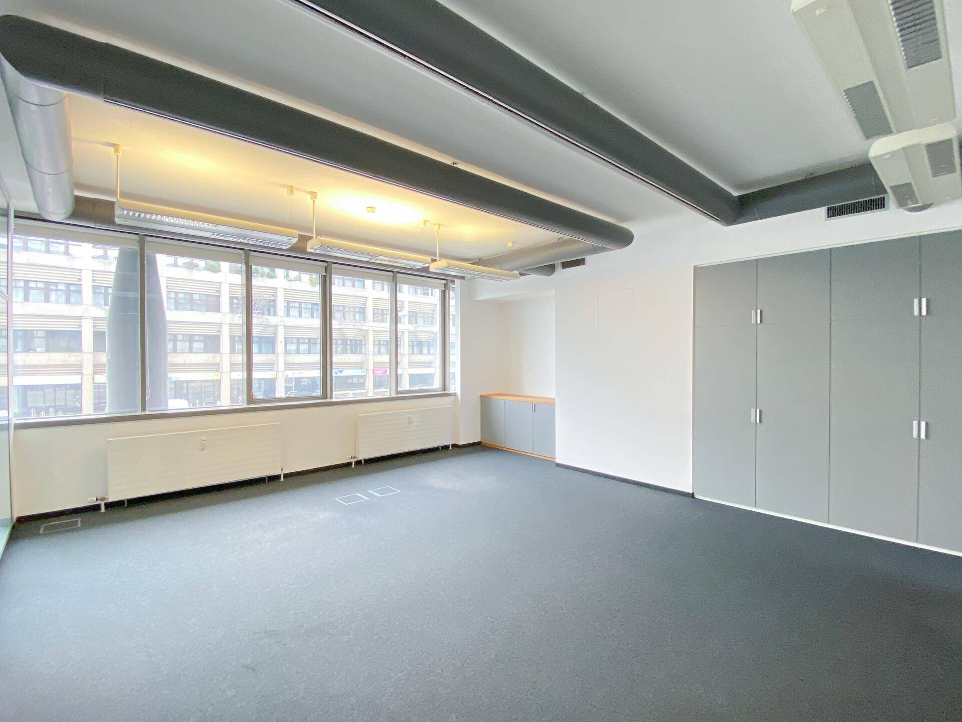 Moderner Büroraum mit Teppichboden, großen Fenstern, sichtbaren Lüftungsrohren und Einbauschränken.