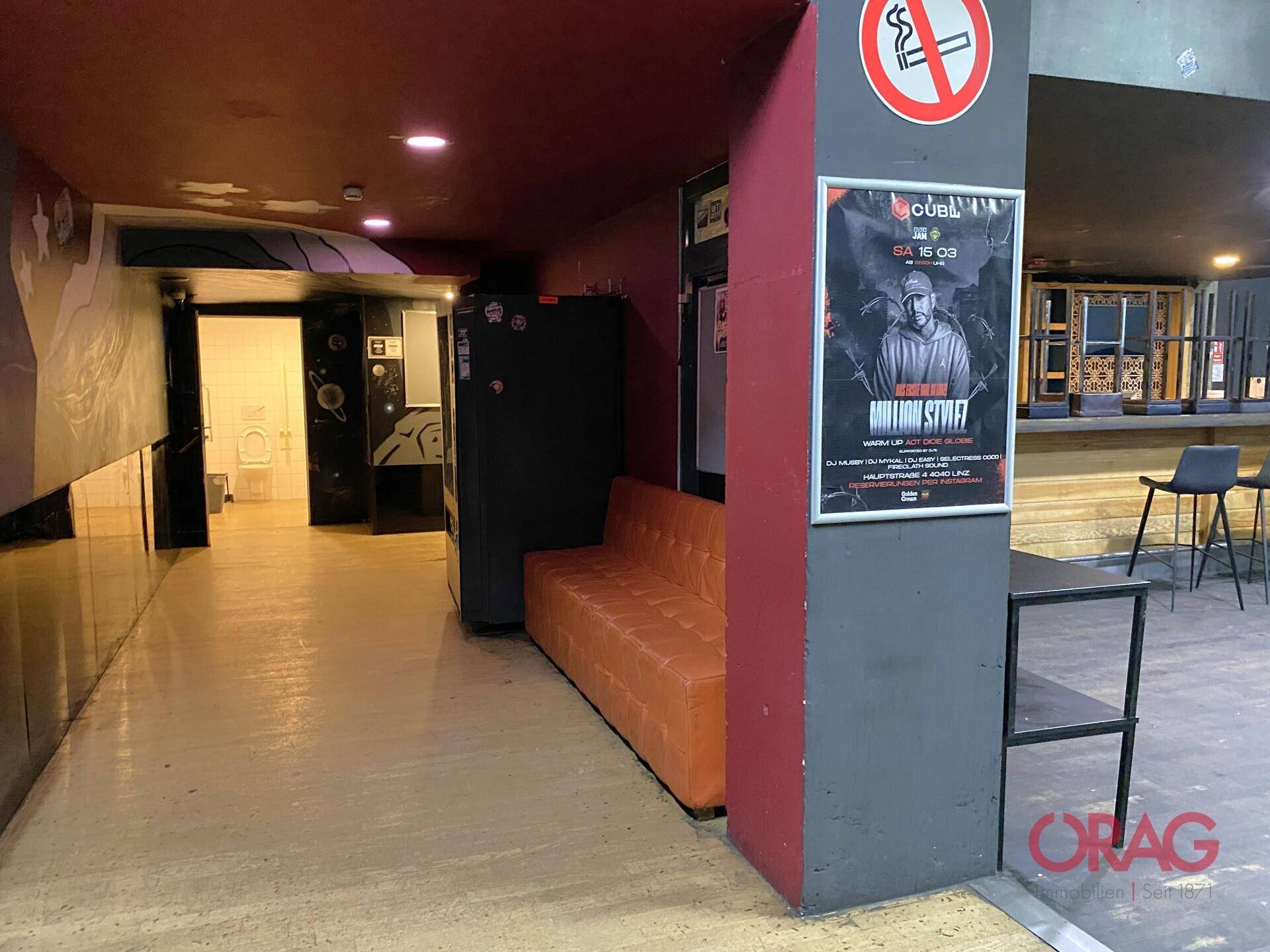 Flurbereich mit orangefarbenem Sofa, Plakaten und Zugang zu weiteren Räumen, mit hellen Böden.