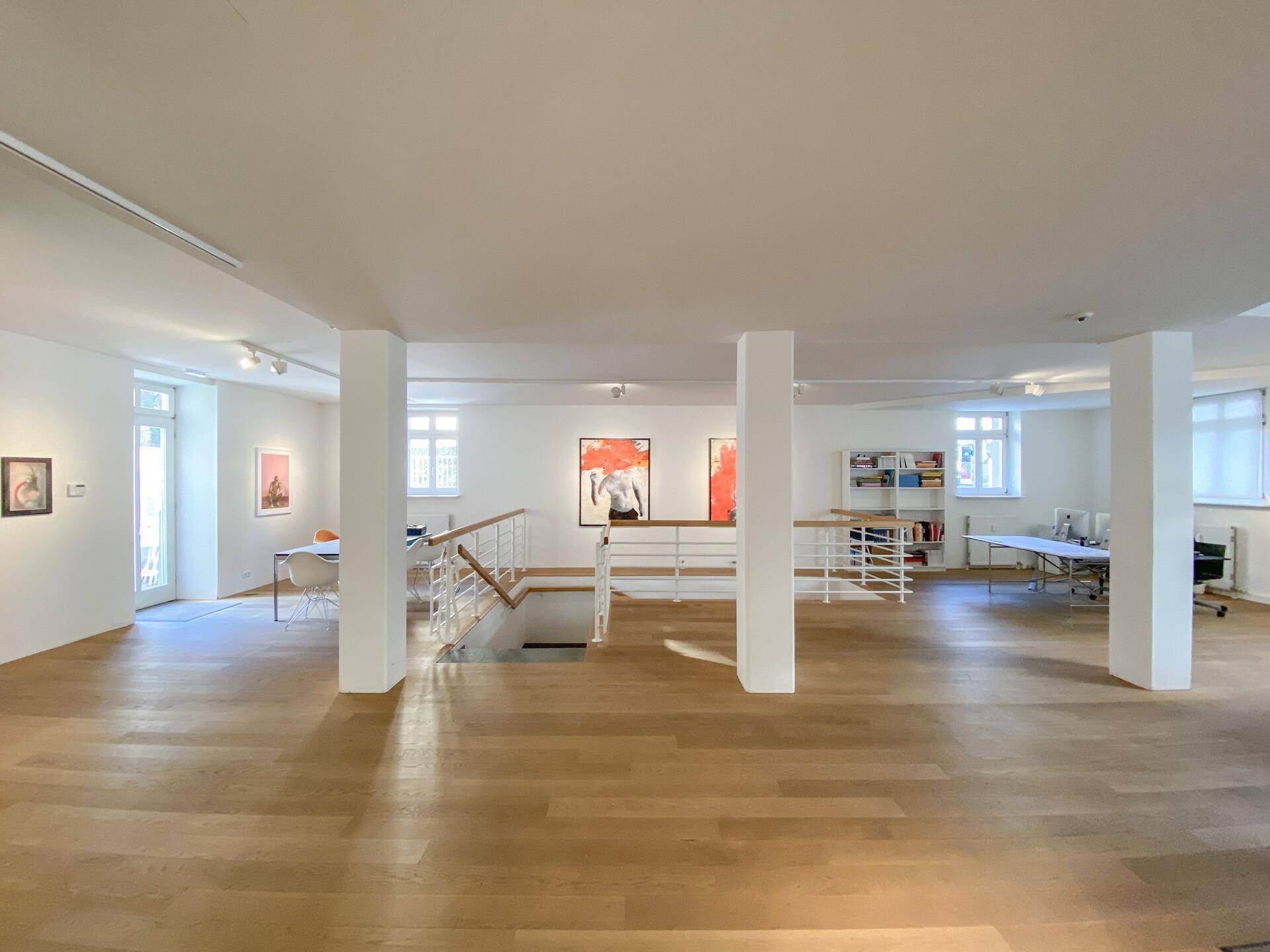 Großer, heller Raum mit Holzboden, der als Ausstellungsfläche und Büro genutzt wird, mit Blick nach draußen.