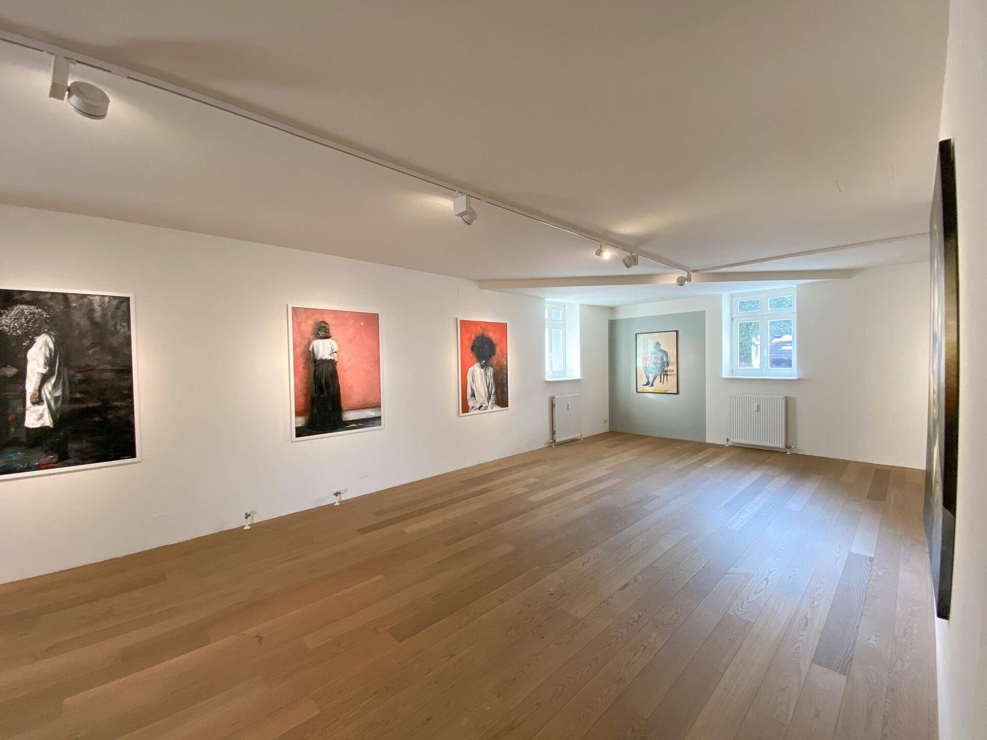 Langer Ausstellungsraum mit Holzboden, Kunstwerken an den Wänden und Fenstern, die Tageslicht hereinlassen.