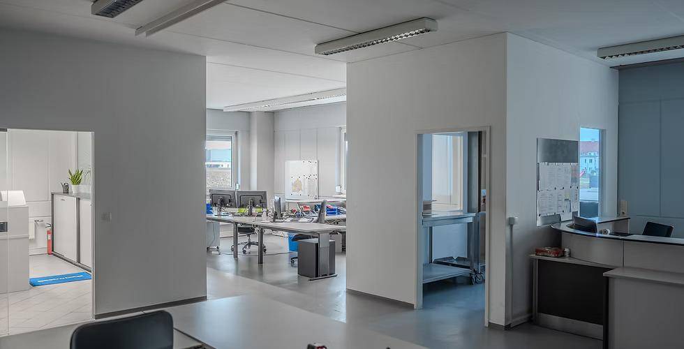 Offener Bürobereich mit mehreren Arbeitsplätzen, hellen Wänden und funktionaler Beleuchtung, ideal für Teamarbeit.