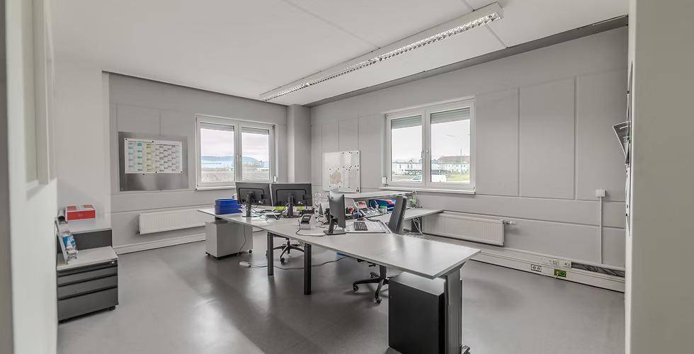 Helles und funktionales Büro mit mehreren Arbeitsplätzen, großen Fenstern und grauem Bodenbelag.