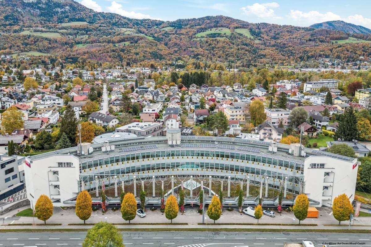 Luftaufnahme eines großen, geschwungenen Bürogebäudes mit Herbstbäumen und Bergen im Hintergrund, umgeben von Wohngebieten.