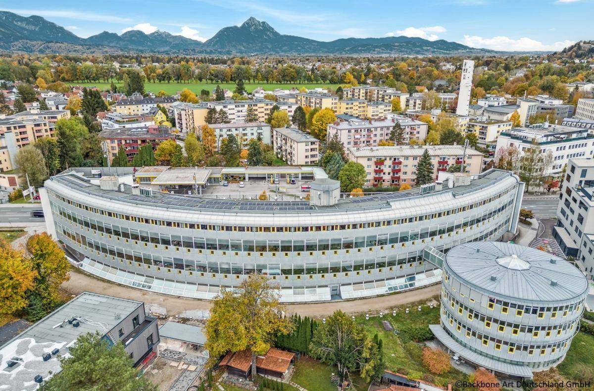 Weitläufige Luftaufnahme des geschwungenen Bürokomplexes mit umliegender Bebauung und Bergpanorama.