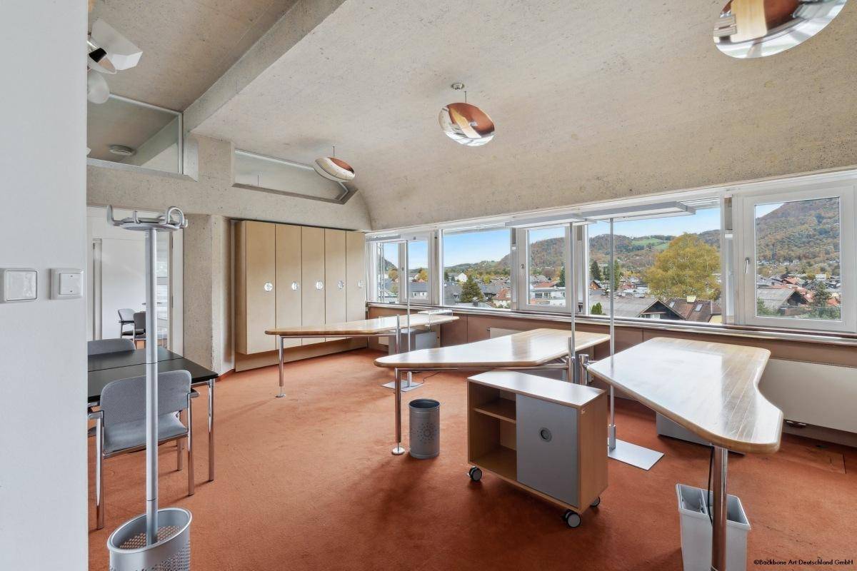 Büro mit mehreren Arbeitsplätzen, Teppichboden und großen Fenstern mit Blick auf die grüne Umgebung.