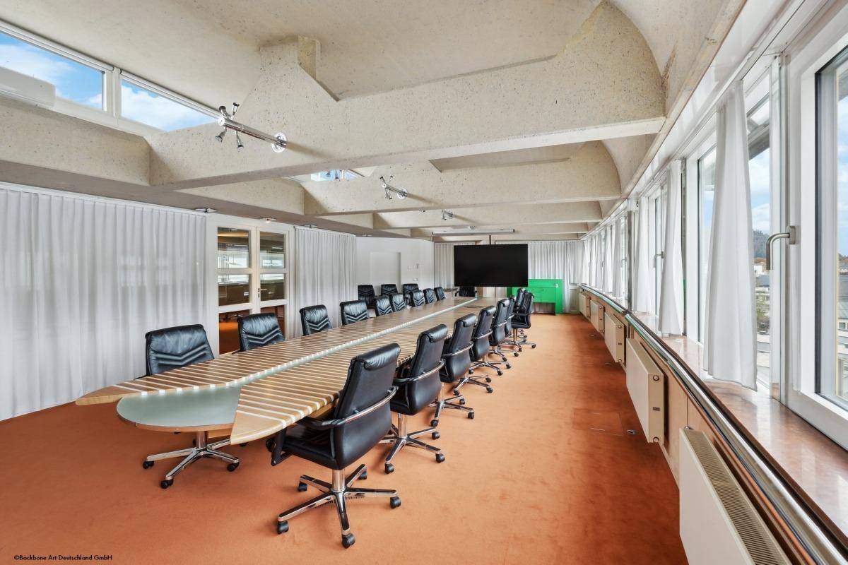 Großer Konferenzraum mit langem Holztisch, vielen Stühlen und einer Fensterfront mit Vorhängen.