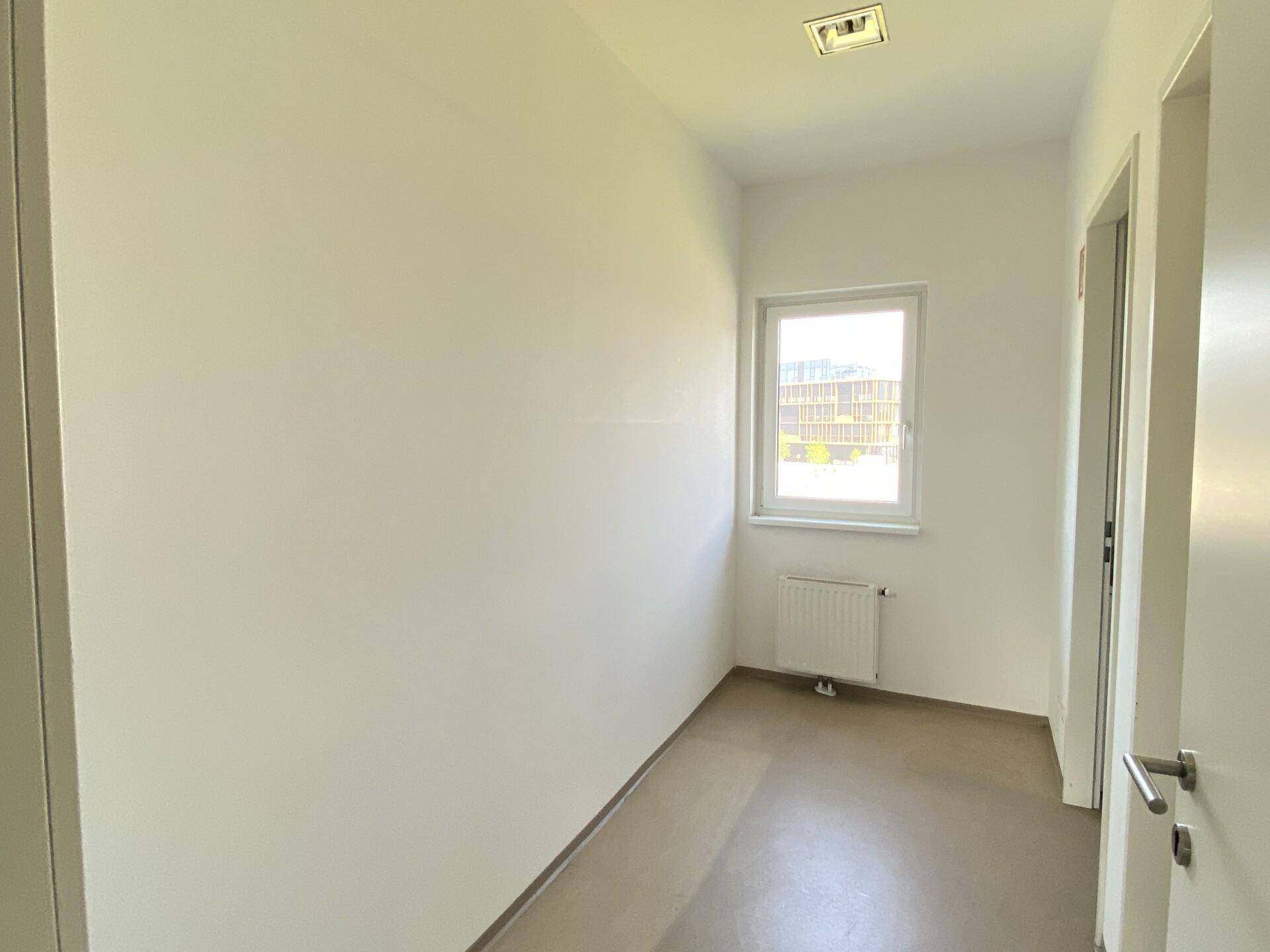 Schmaler Flur mit Fenster, weißen Wänden und dunklem Boden, bietet natürlichen Lichteinfall.