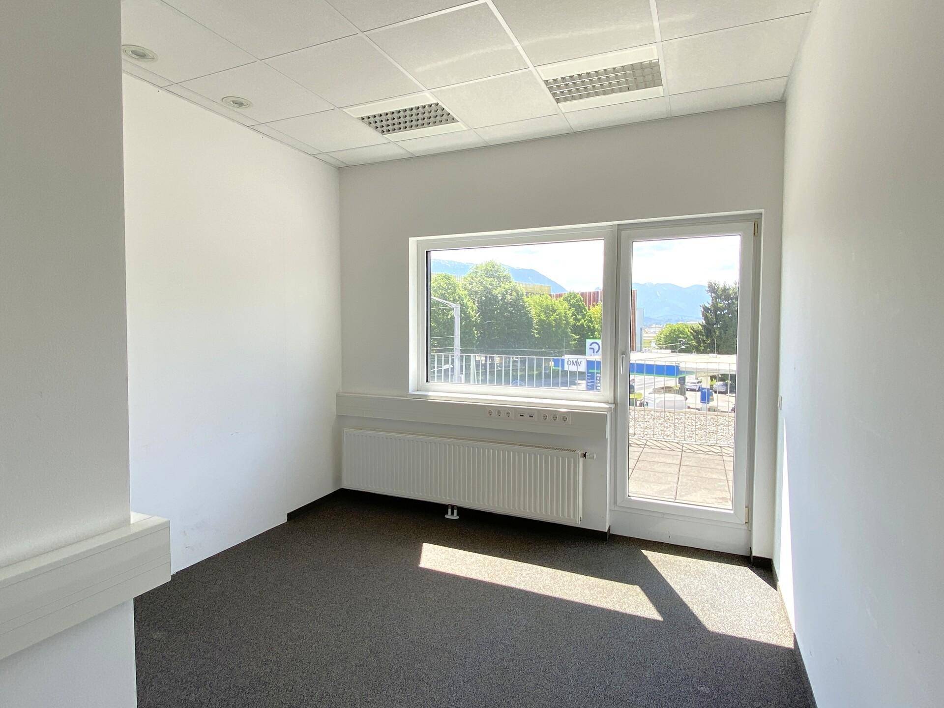 Kleines Büro mit Teppichboden, Fenster und Glastür, bietet Blick auf die Straße und entfernte Berge.