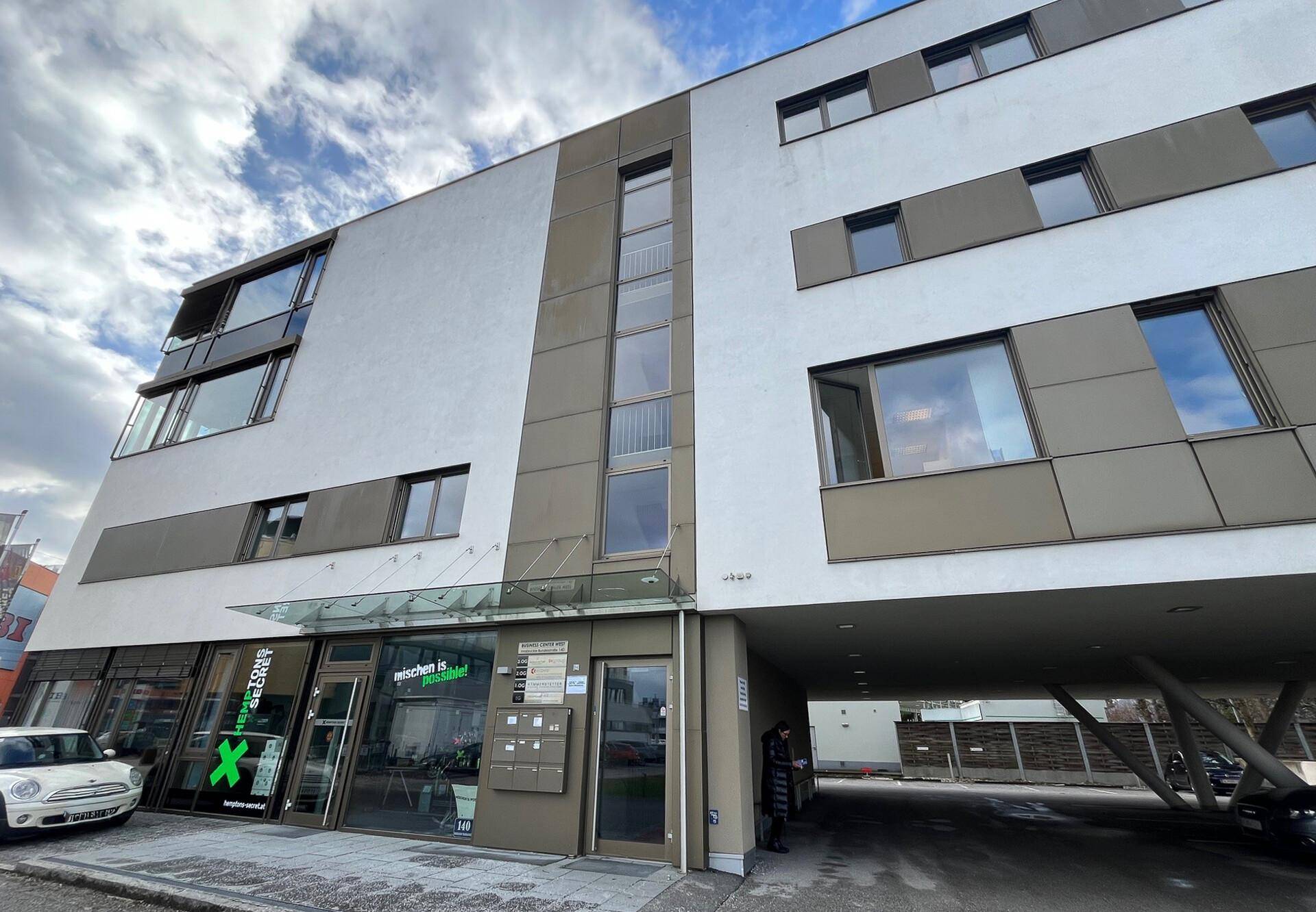 Moderne Geschäfts- und Wohngebäude-Fassade mit großen Fenstern und überdachtem Parkbereich.