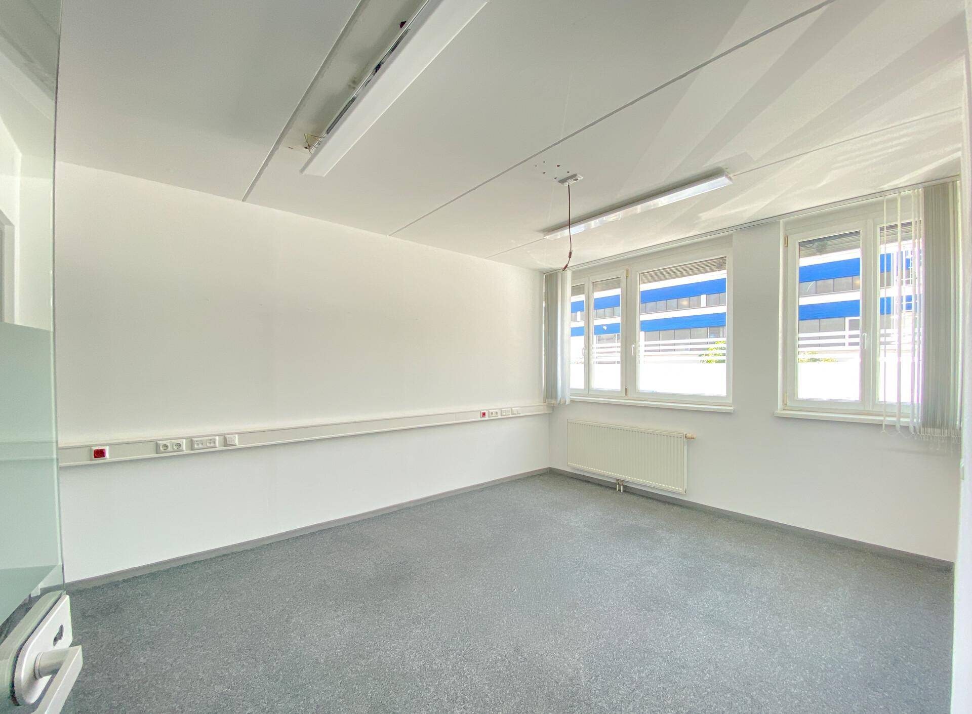 Helles Büro mit Teppichboden, Heizkörper und Fenstern mit Jalousien für angenehmes Licht.