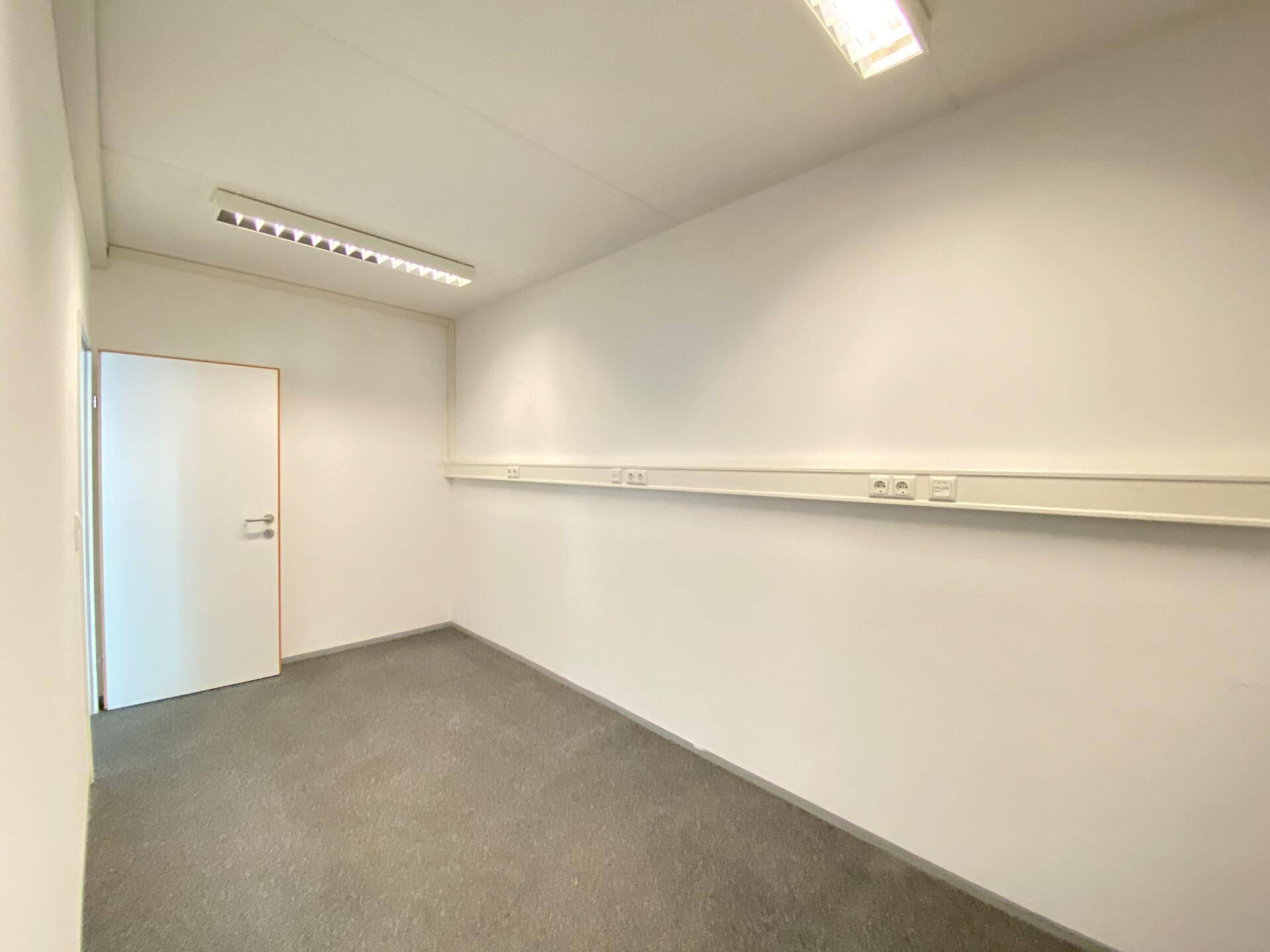 Kleines, helles Büro mit Teppichboden, weißen Wänden und praktischen Wandsteckdosenleisten.