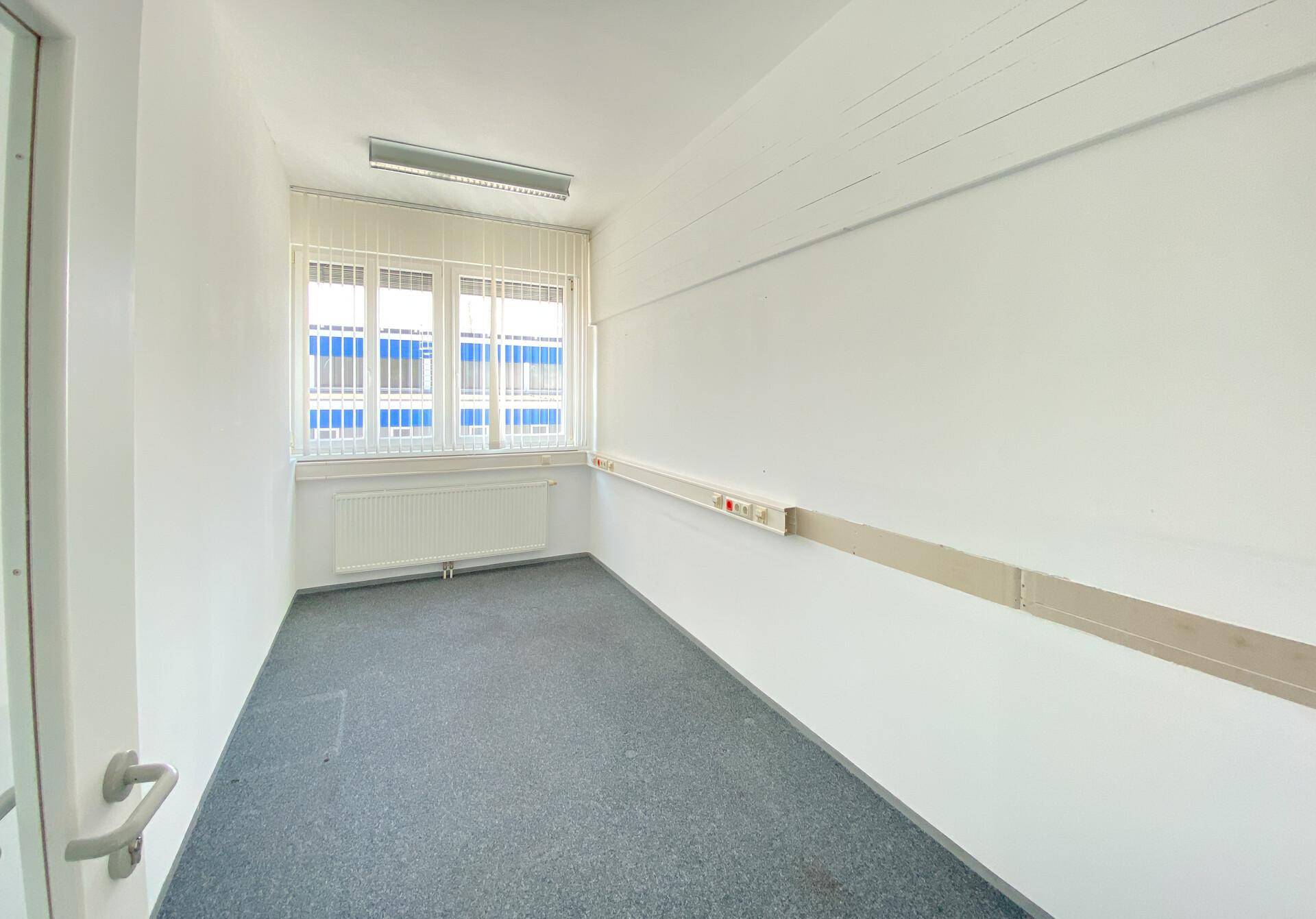 Kleines Büro mit Teppichboden, Fenster mit Jalousien, Heizkörper und einer Glastür.