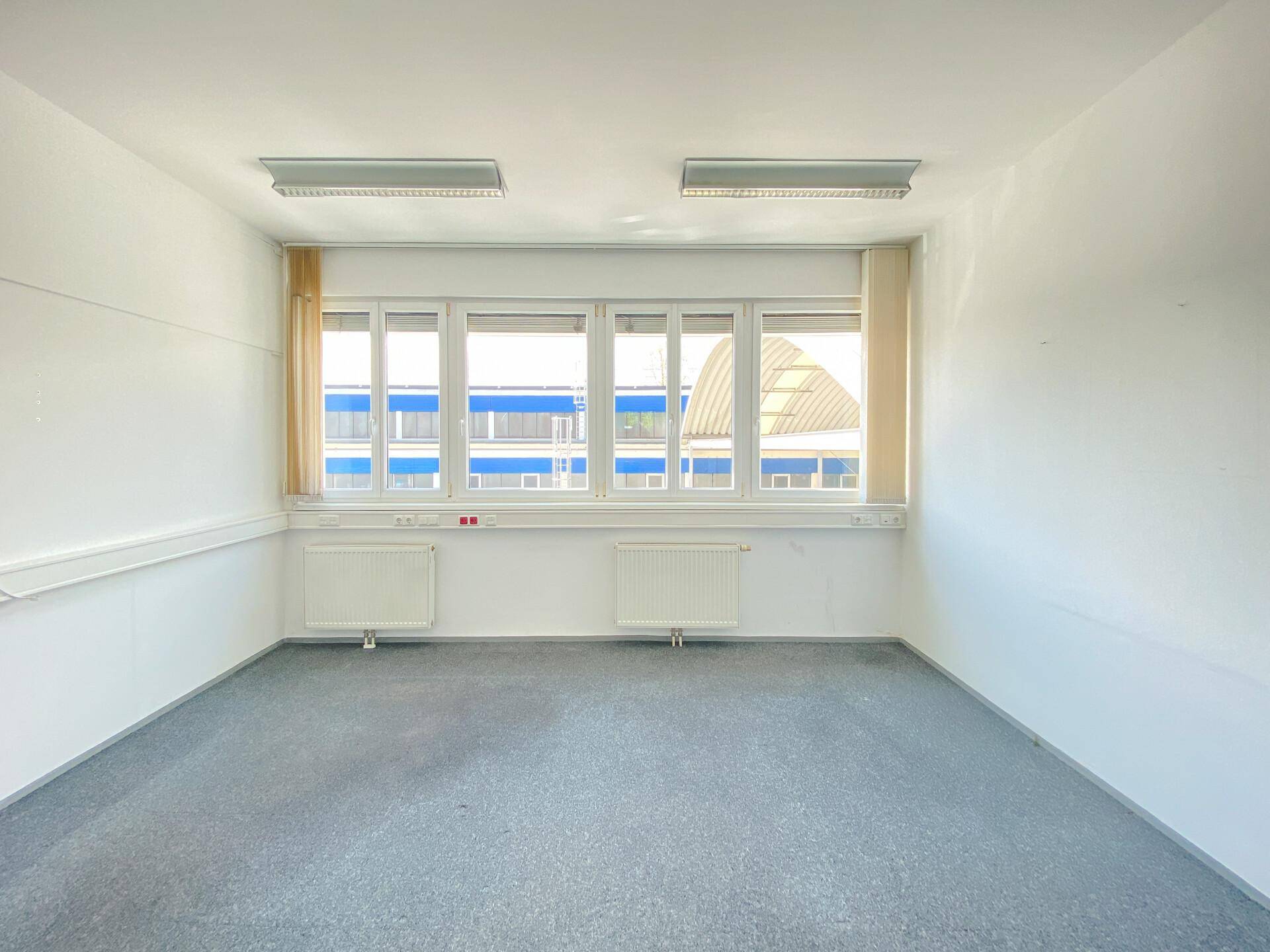 Helles Büro mit Teppichboden, Heizkörpern und großen Fenstern mit Jalousien für Privatsphäre.
