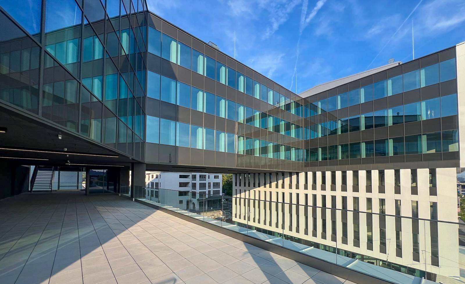 Eine weitläufige Terrasse mit Glasgeländer bietet Blick auf die moderne Gebäudefassade und den Himmel.